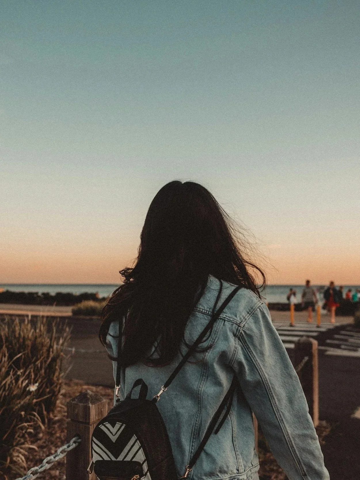 Person with long dark hair wearing a denim jacket carrying a small black and white backpack, standing near a coastal area at sunset with a group of people in the distance.