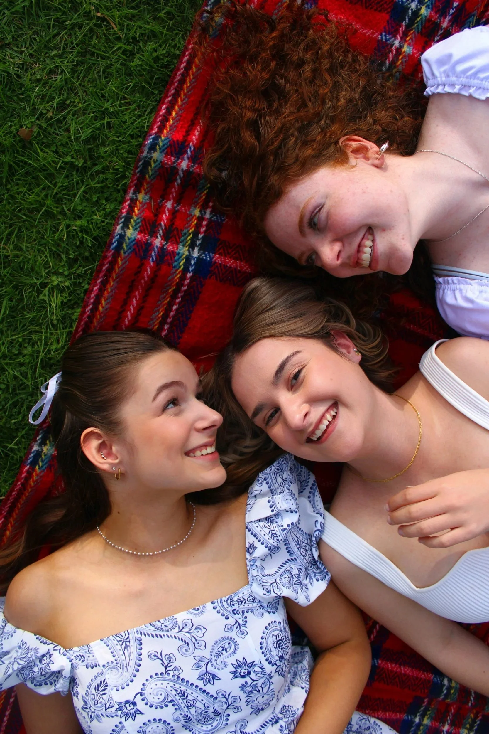 Three young women lying on a red and blue plaid blanket on green grass, smiling and looking at each other.