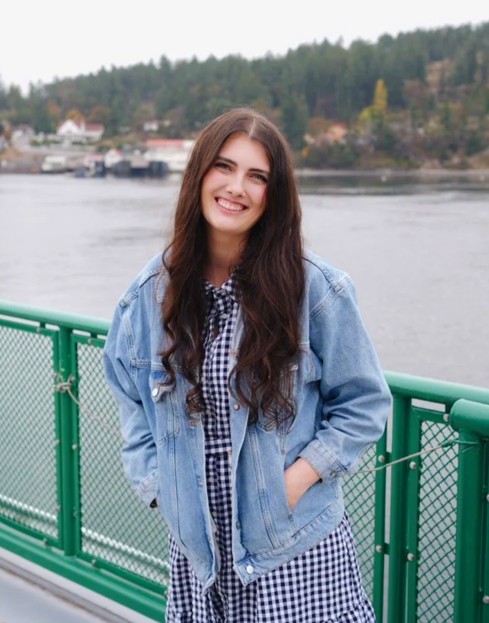 A young woman with long brown hair smiling, wearing a denim jacket over a black and white gingham dress, standing on a boat or ferry with water and trees in the background.