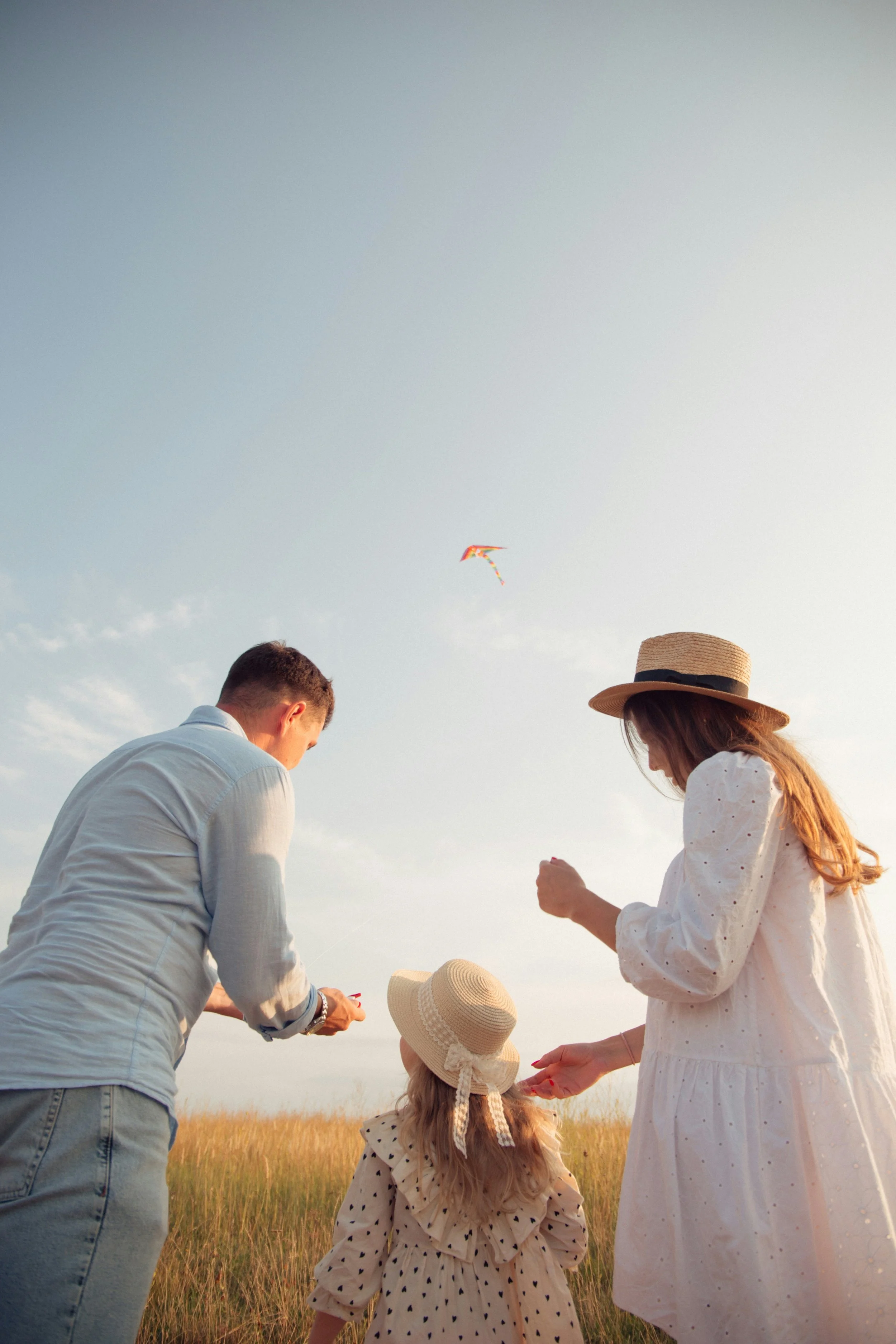 A family of three flying a kite in a grassy field during sunset. The family includes a man, a woman, and a young girl wearing a hat. The sky is partly cloudy with a colorful kite flying high.