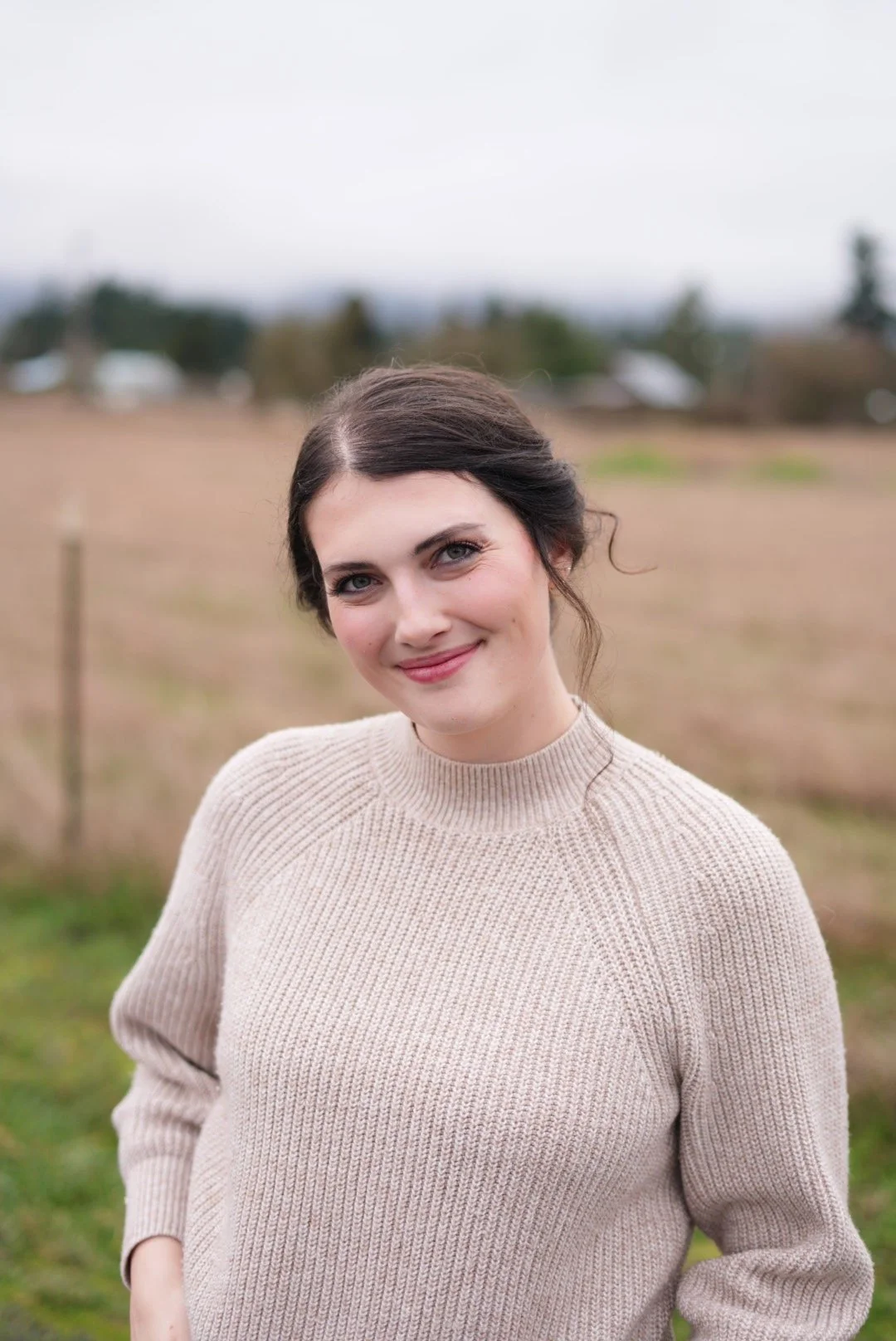 A young woman with dark hair styled in a loose bun, wearing a beige knit sweater, smiling outdoors in a field with a cloudy sky in the background.