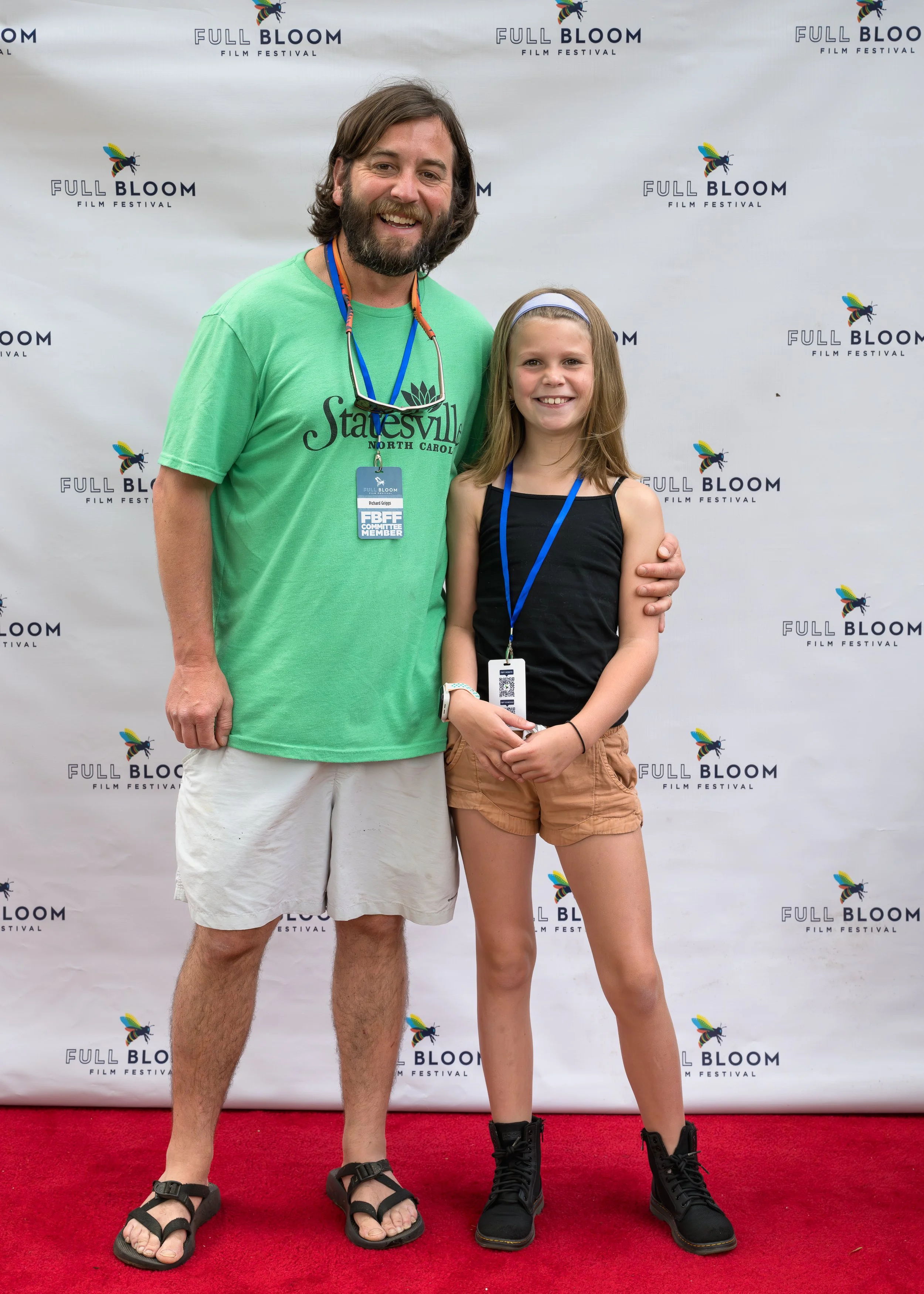 Man and young girl standing together on a red carpet at the Full Bloom Film Festival, smiling at the camera. The man is wearing a green t-shirt with a logo, beige shorts, and sandals, while the girl is in a black tank top, tan shorts, and black boots. Both have event badges on blue lanyards around their necks.