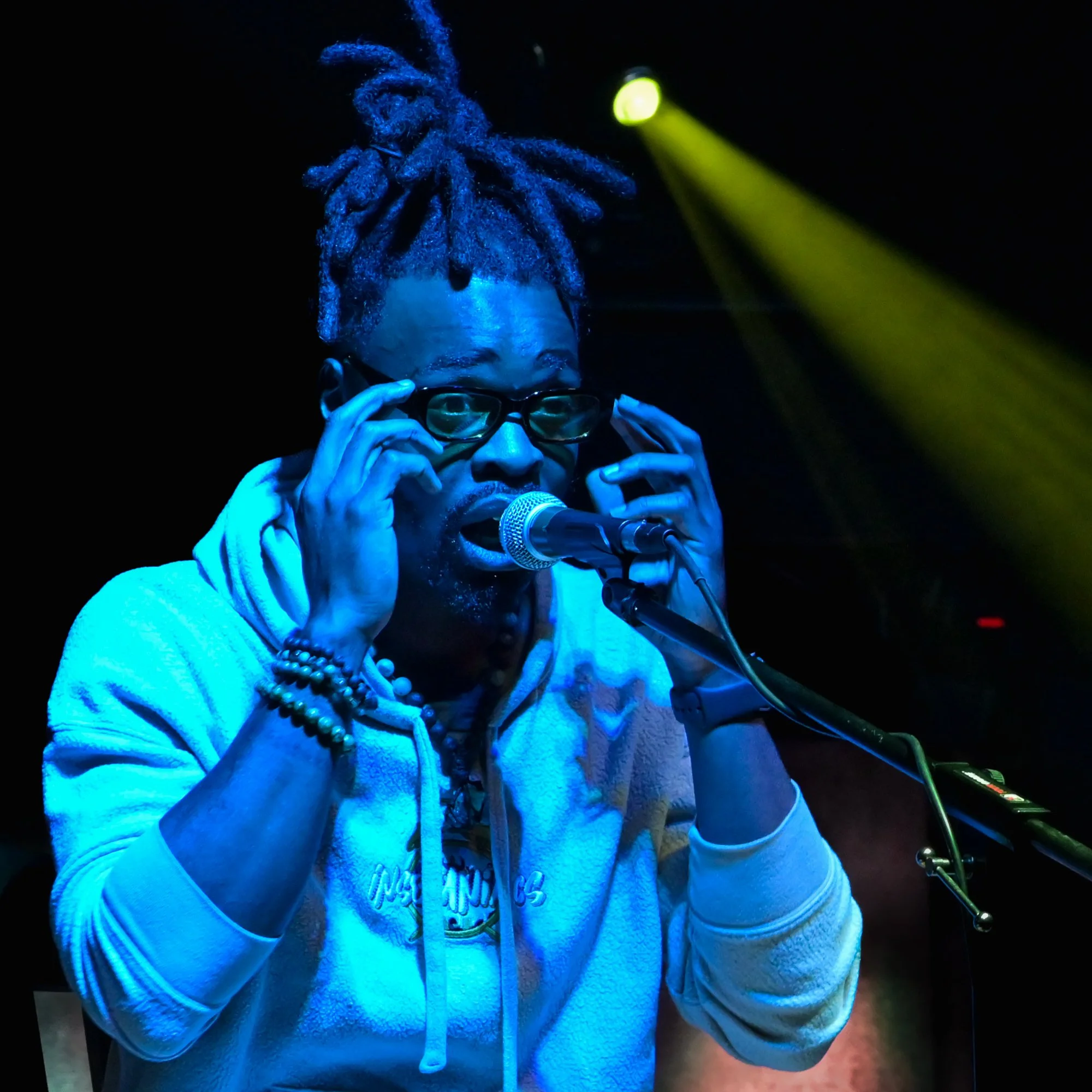 A man with dreadlocks, glasses, and a hoodie singing into a microphone on stage with colorful lighting.