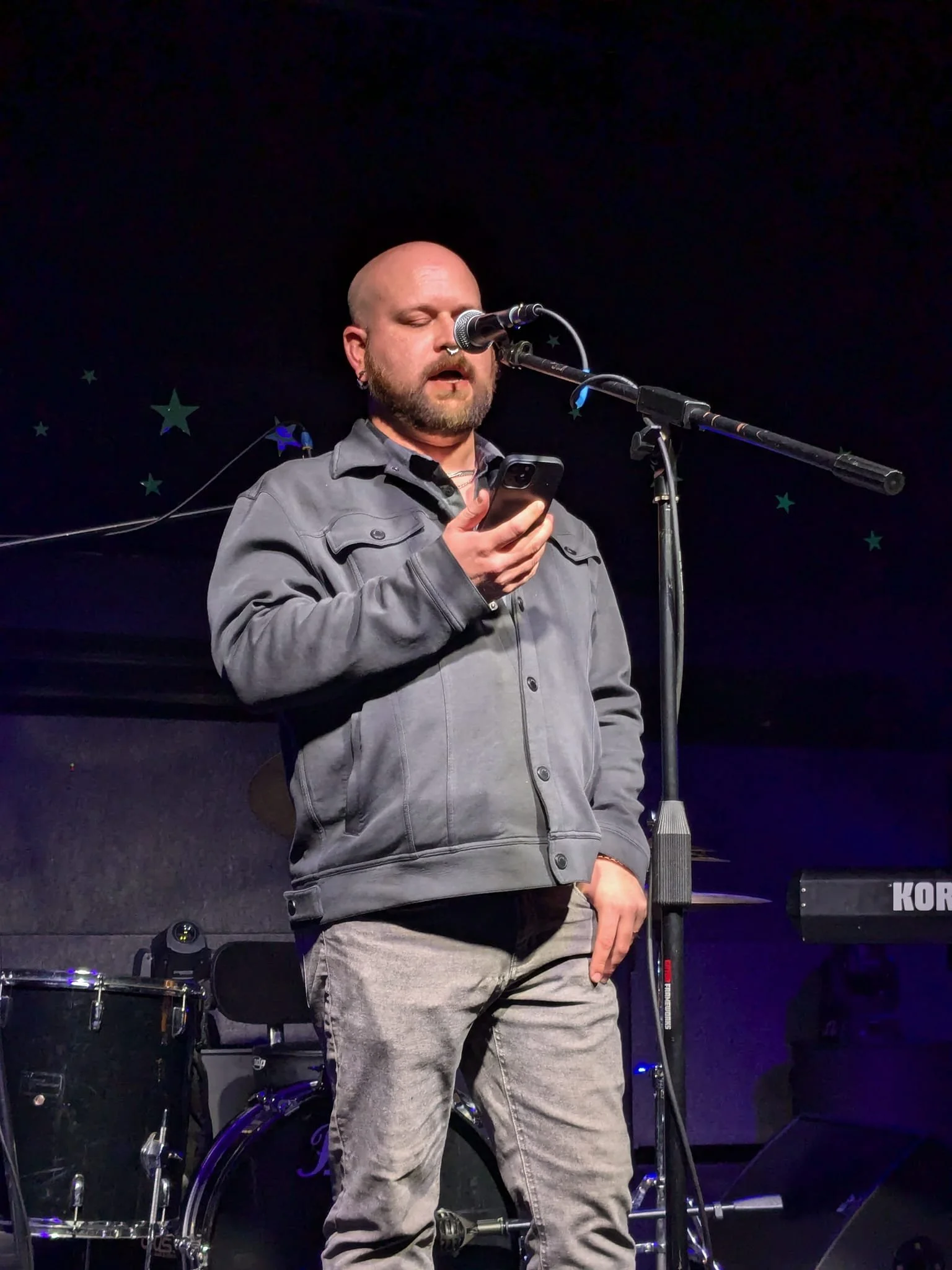 A man with a beard and bald head, wearing a gray jacket and jeans, stands on stage reading from his phone in front of a microphone. Musical instruments are visible in the background.