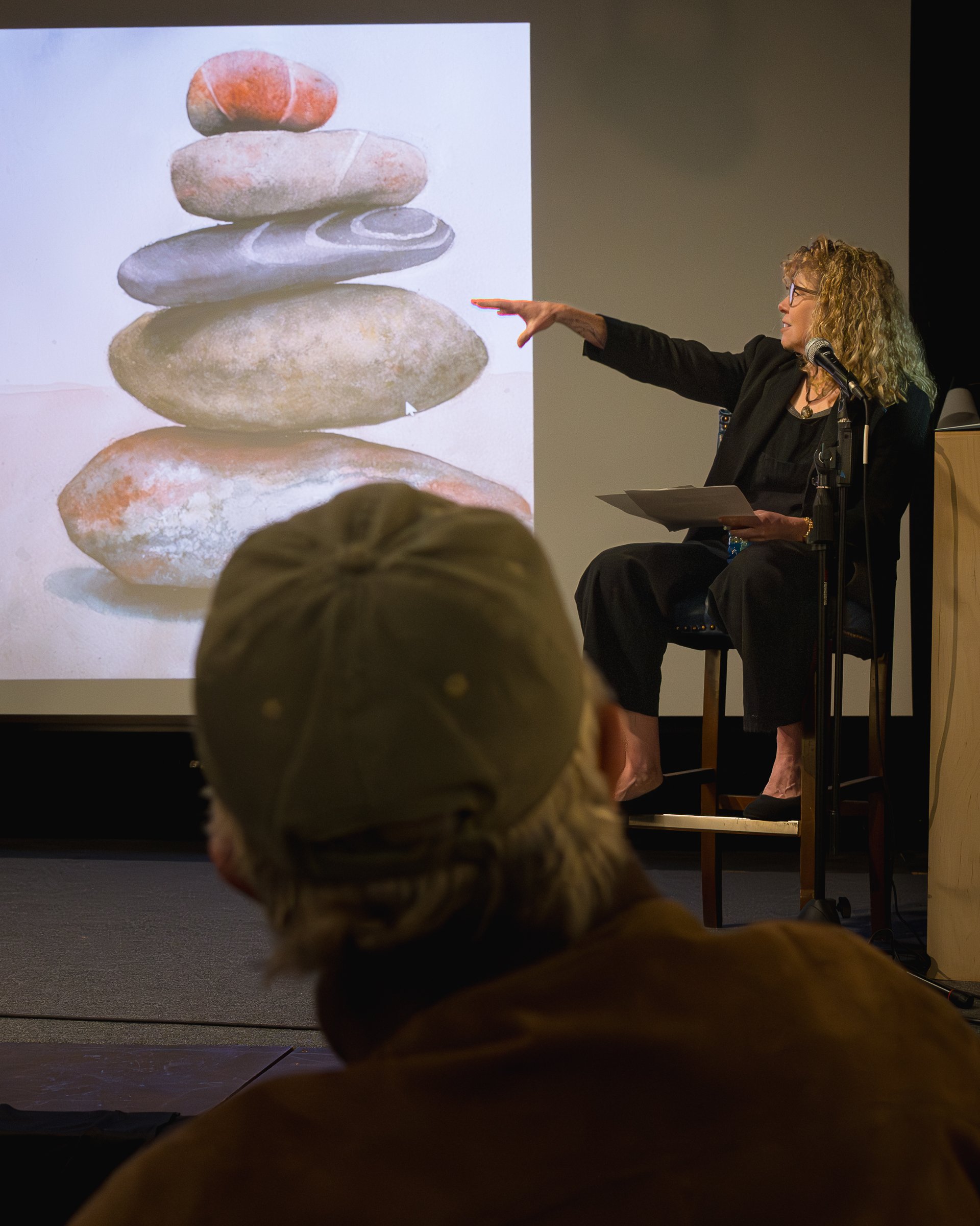A woman with curly blonde hair and glasses is seated on a stool, pointing towards a projected image of stacked rocks during a presentation.