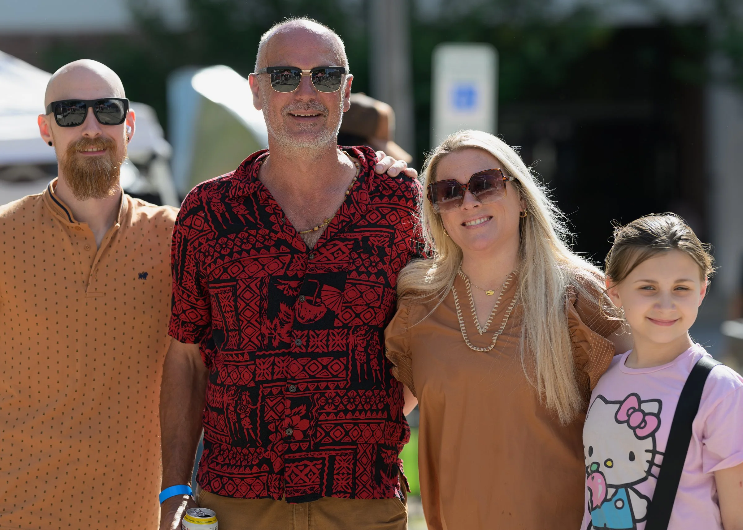 A group of four people smiling outdoors on a sunny day, including a man with a shaved head and sunglasses, a woman with long blonde hair and sunglasses, a young girl with brown hair and a Hello Kitty shirt, and a man with a beard and sunglasses.