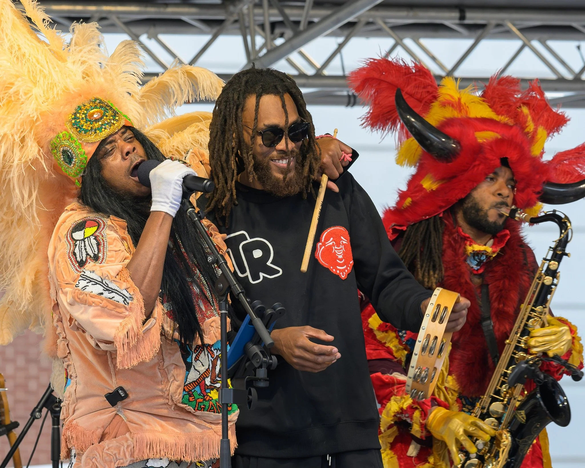 Musicians performing on stage, dressed in vibrant, elaborate costumes including feathered headdresses and animal-themed outfits, with one singing into a microphone, another holding a tambourine, and a third playing a saxophone.