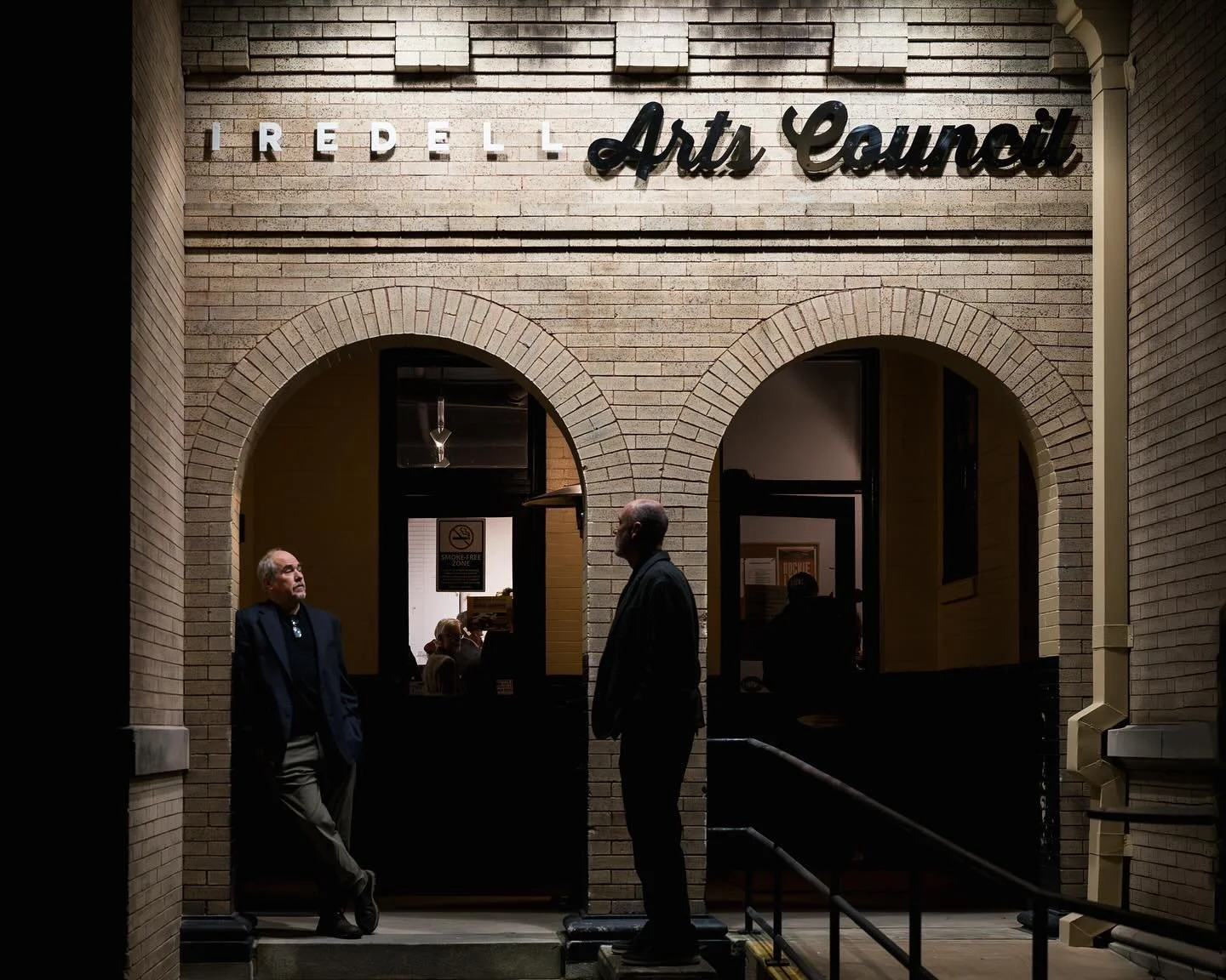 People standing and talking outside the Iredell Arts Council building at night. The building has brick walls and arched windows, with the sign 'Iredell Arts Council' above.