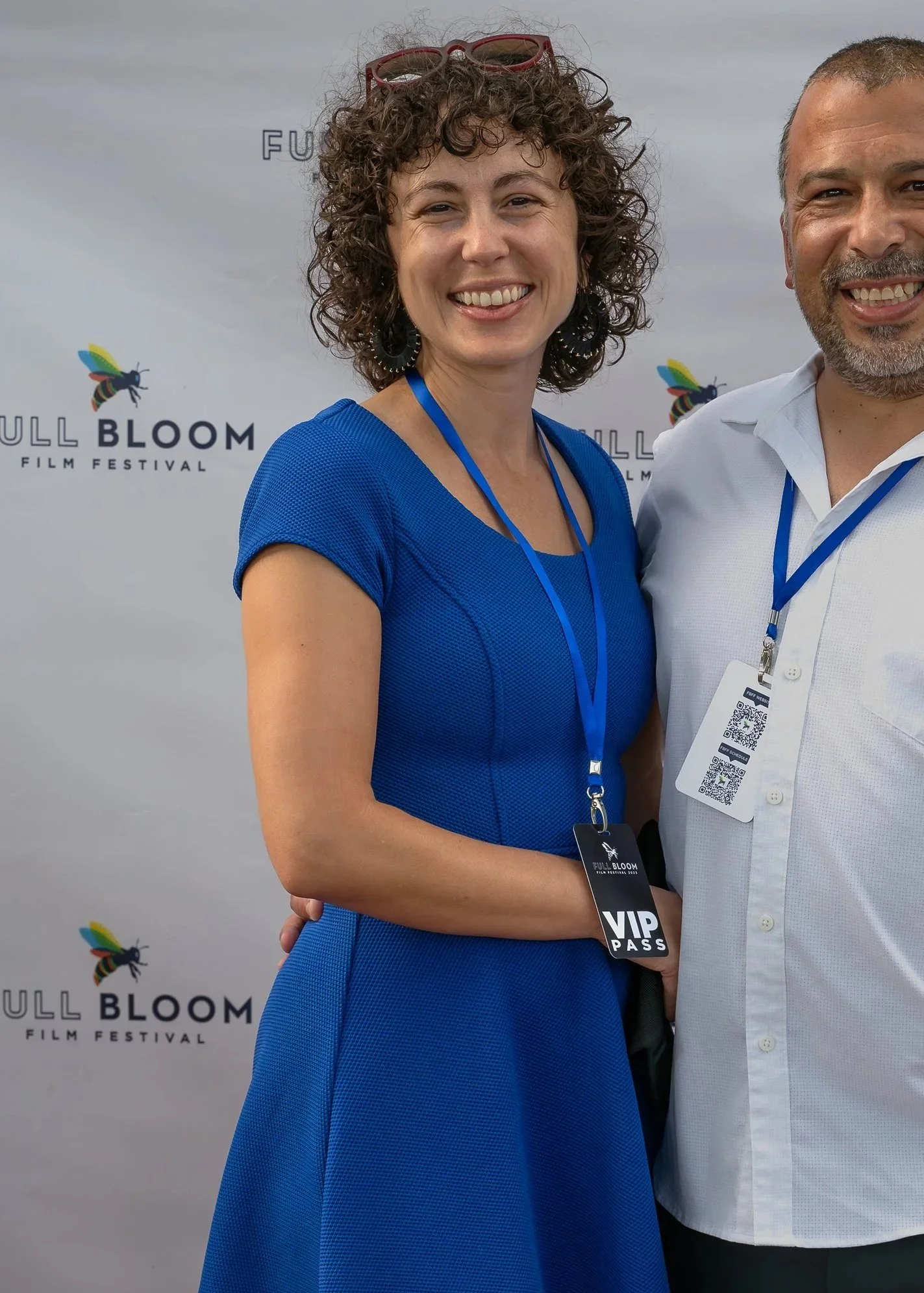 A woman with curly hair wearing a blue dress and red sunglasses on her head standing next to a man in a white shirt at the Full Bloom Film Festival. Both are wearing VIP passes around their necks and smiling for the photo.