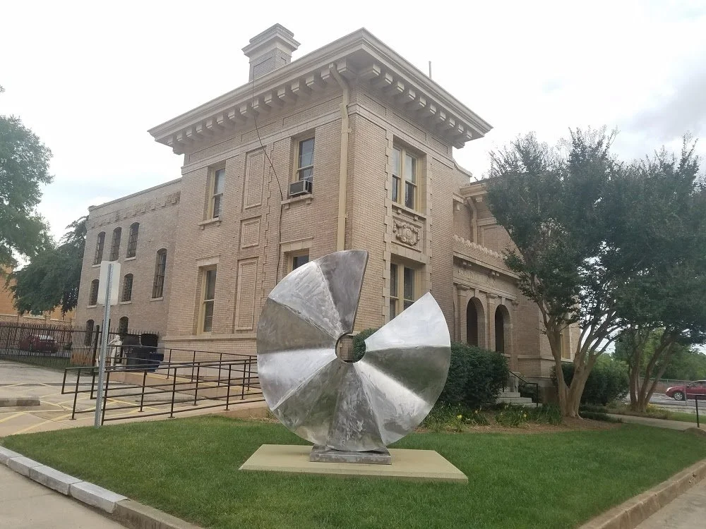 A modern abstract metal sculpture with curved, shiny surfaces and a central hole, situated on a small concrete pad in front of a historic brick building with decorative architectural details and trees.