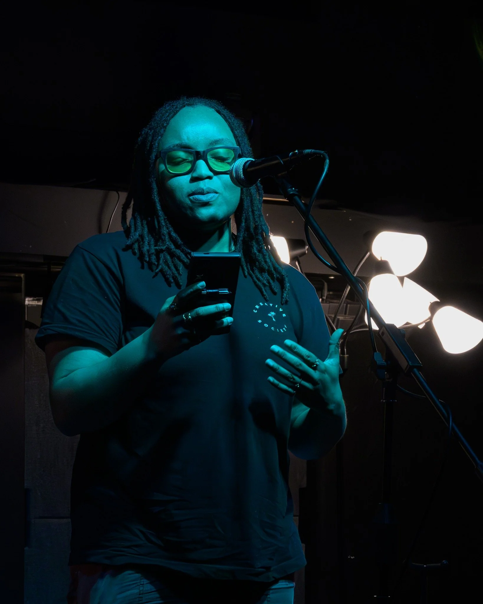 A woman with glasses and dreadlocks performing on stage, holding a phone near her face and standing in front of a microphone with stage lights behind her.