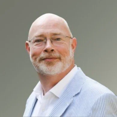 A middle-aged man with glasses, a beard, and a bald head, wearing a white shirt and a light-striped blazer, smiling at the camera.
