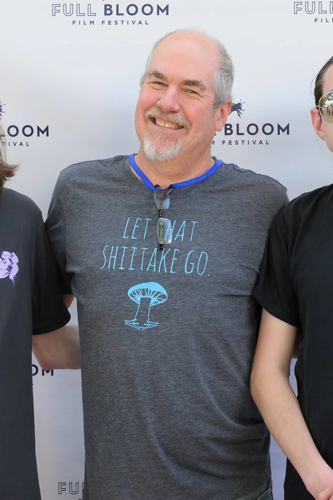 A man smiling at an event, wearing a dark gray T-shirt with a blue mushroom graphic and the phrase "Let That ShiiTake Go." printed on it. He has glasses hanging from his neck and a light beard and mustache. The background features a step-and-repeat banner with the words "Full Bloom Film Festival" and small bug illustrations.