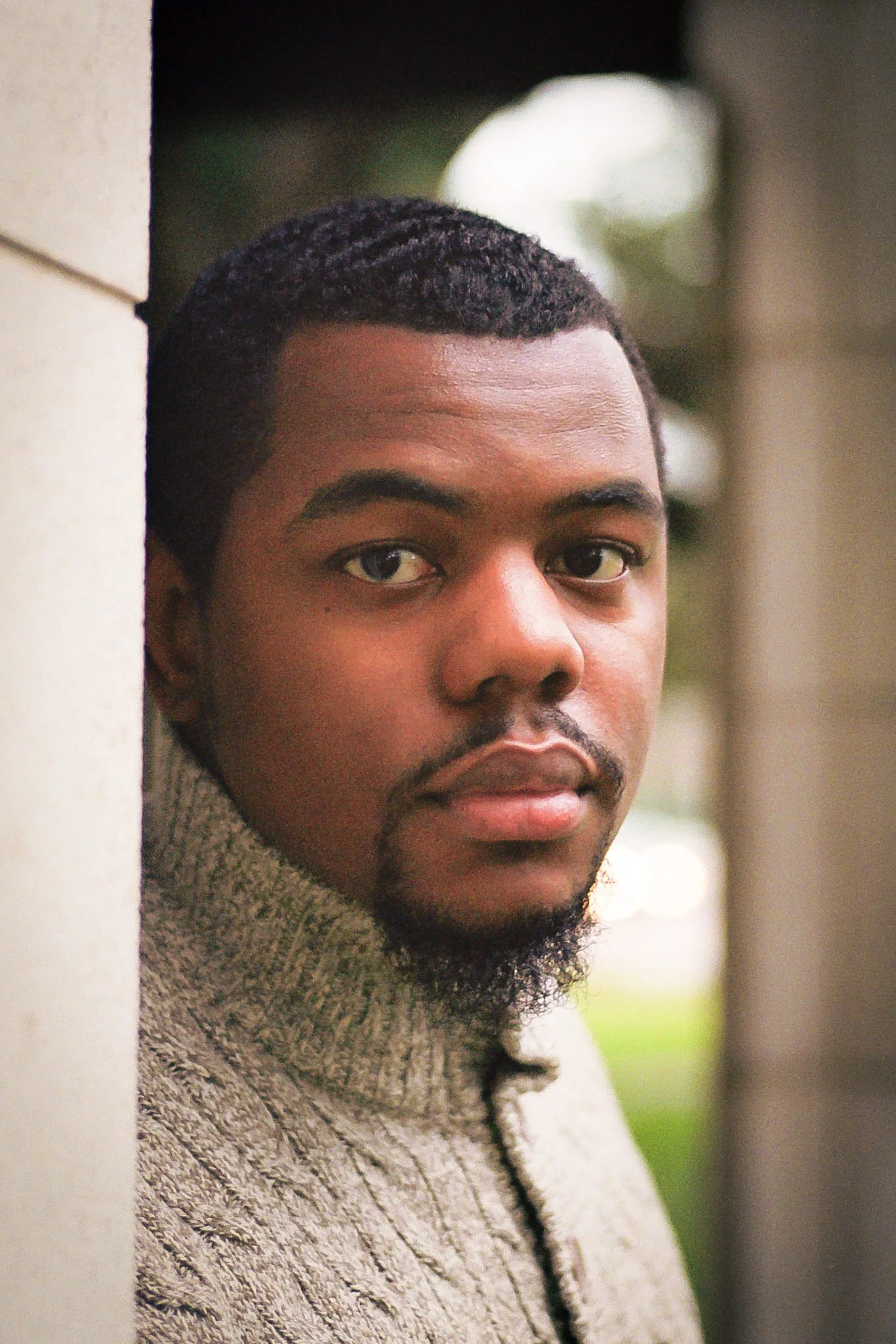 Close-up of a young man with dark hair and beard, leaning against a wall outdoors, looking at the camera.