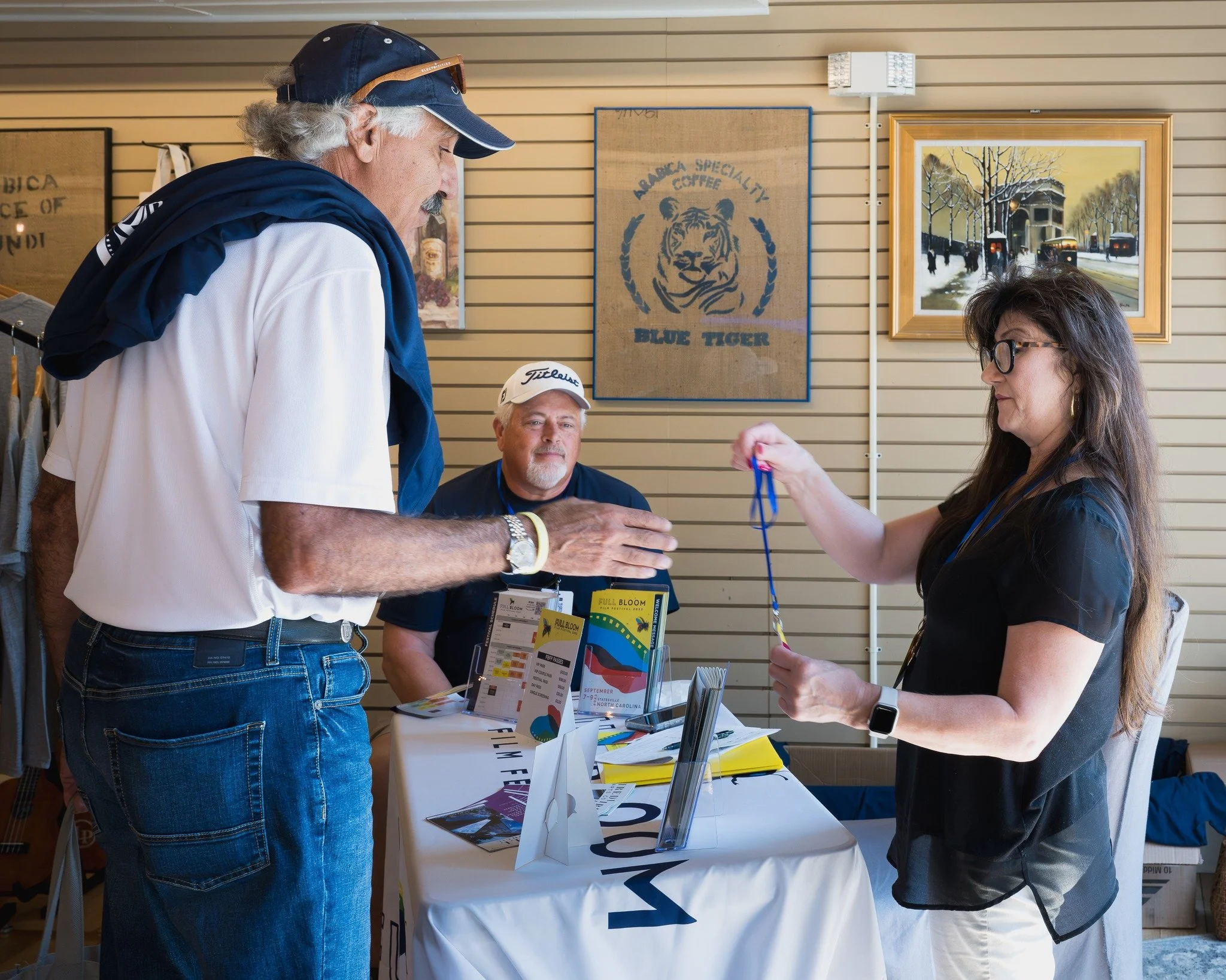 A woman in black with glasses handing a medal to an older man in a white shirt and blue cap at a booth, with another man sitting behind the table. The booth has informational pamphlets and posters, with artwork on the wall behind.