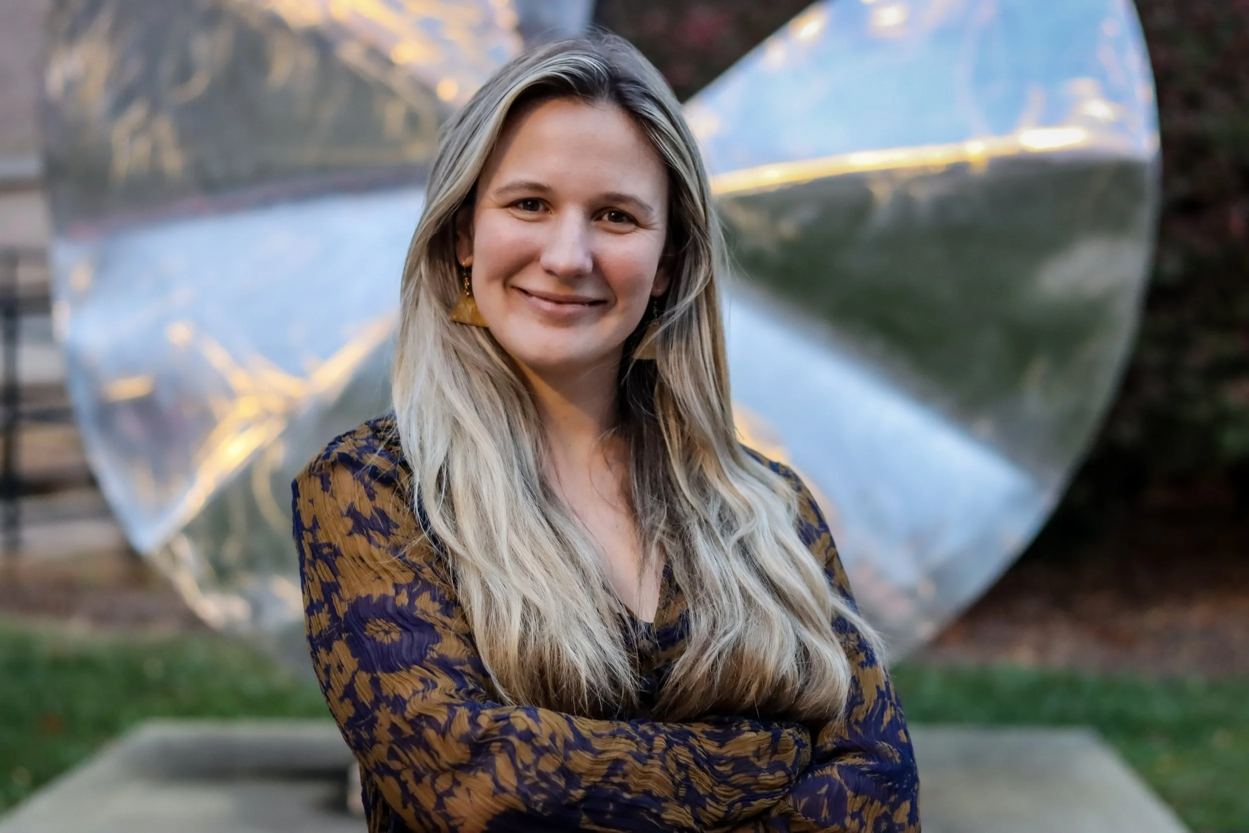 A woman with long blonde hair wearing a patterned top, smiling with her arms crossed, standing outdoors in front of a reflective metallic sculpture.