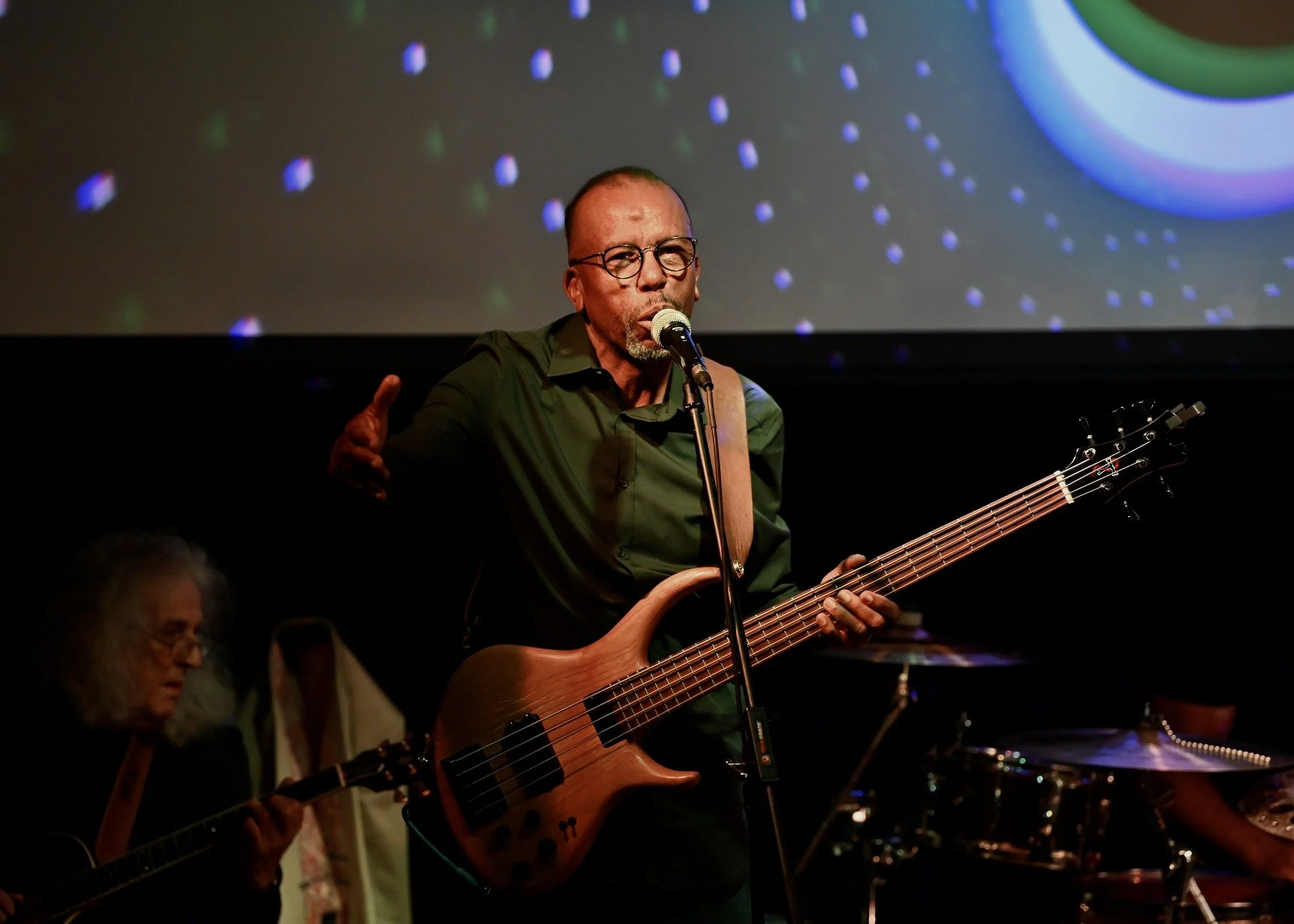 A man with glasses and a green shirt is singing into a microphone while playing a bass guitar on stage. A drummer and another guitarist are visible in the background, and a colorful abstract display is projected behind him.