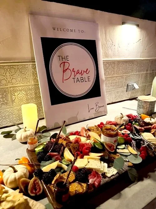 Decorative cheese and fruit platter at a welcoming event named 'The Brave Table' with a sign and candles.