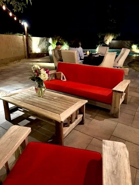 Outdoor patio with wooden furniture, a red sofa, and a table with a vase of flowers, lit by string lights at night.