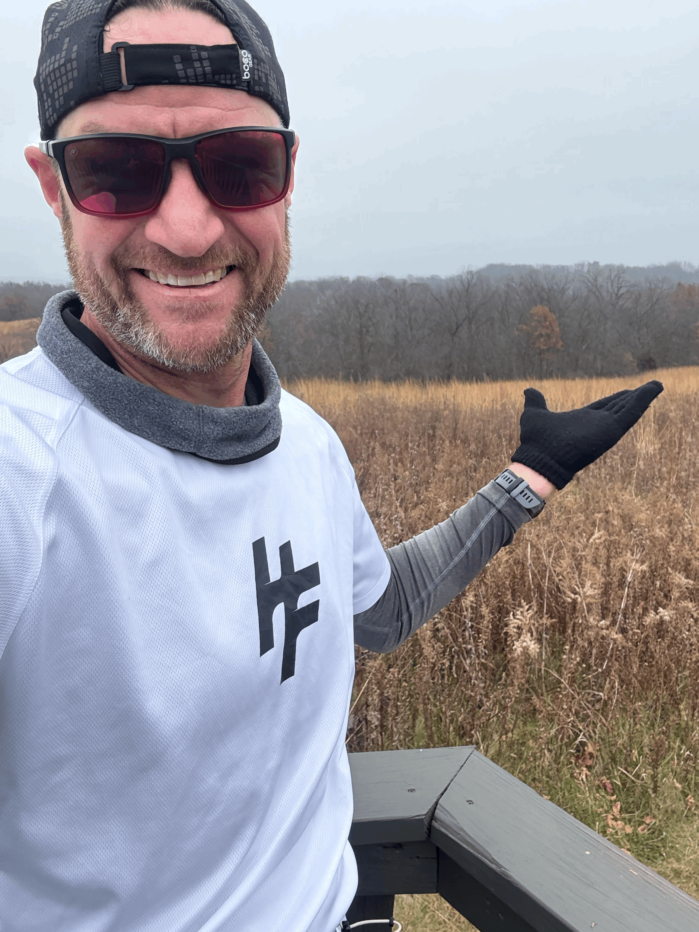A man taking a selfie outdoors on a cloudy day, wearing sunglasses, a black baseball cap, a white sports shirt, and a gray long-sleeve shirt underneath, standing on a wooden platform with dry grass and trees in the background.