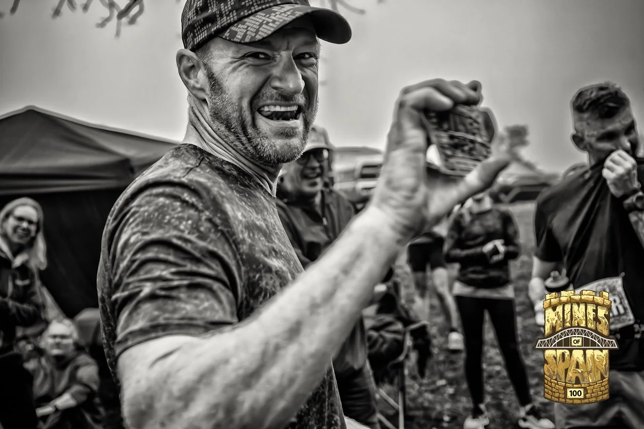 Man smiling and holding a medal or badge, with a group of people in the background, outdoor setting, black and white photo.