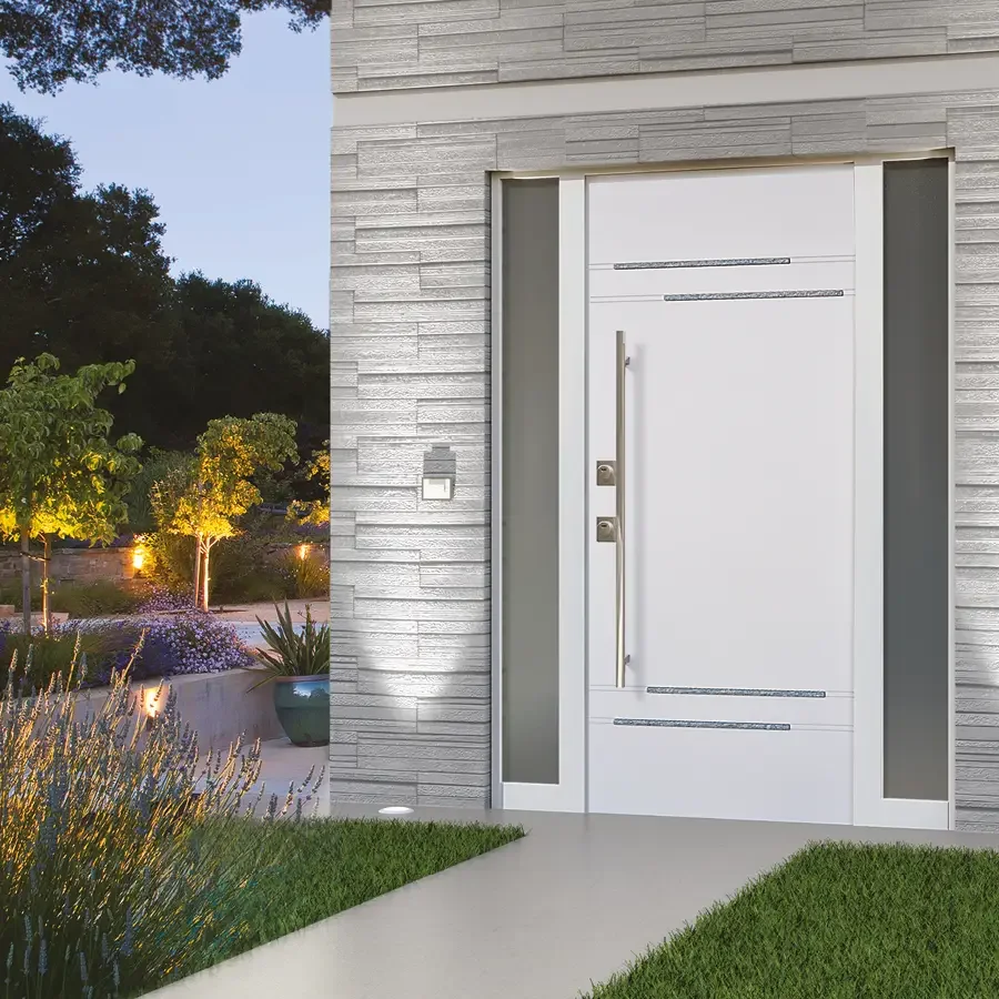 Porta d'ingresso moderna bianca con maniglia lunga davanti a una parete in pietra grigia, con un giardino e alberi illuminati sullo sfondo.