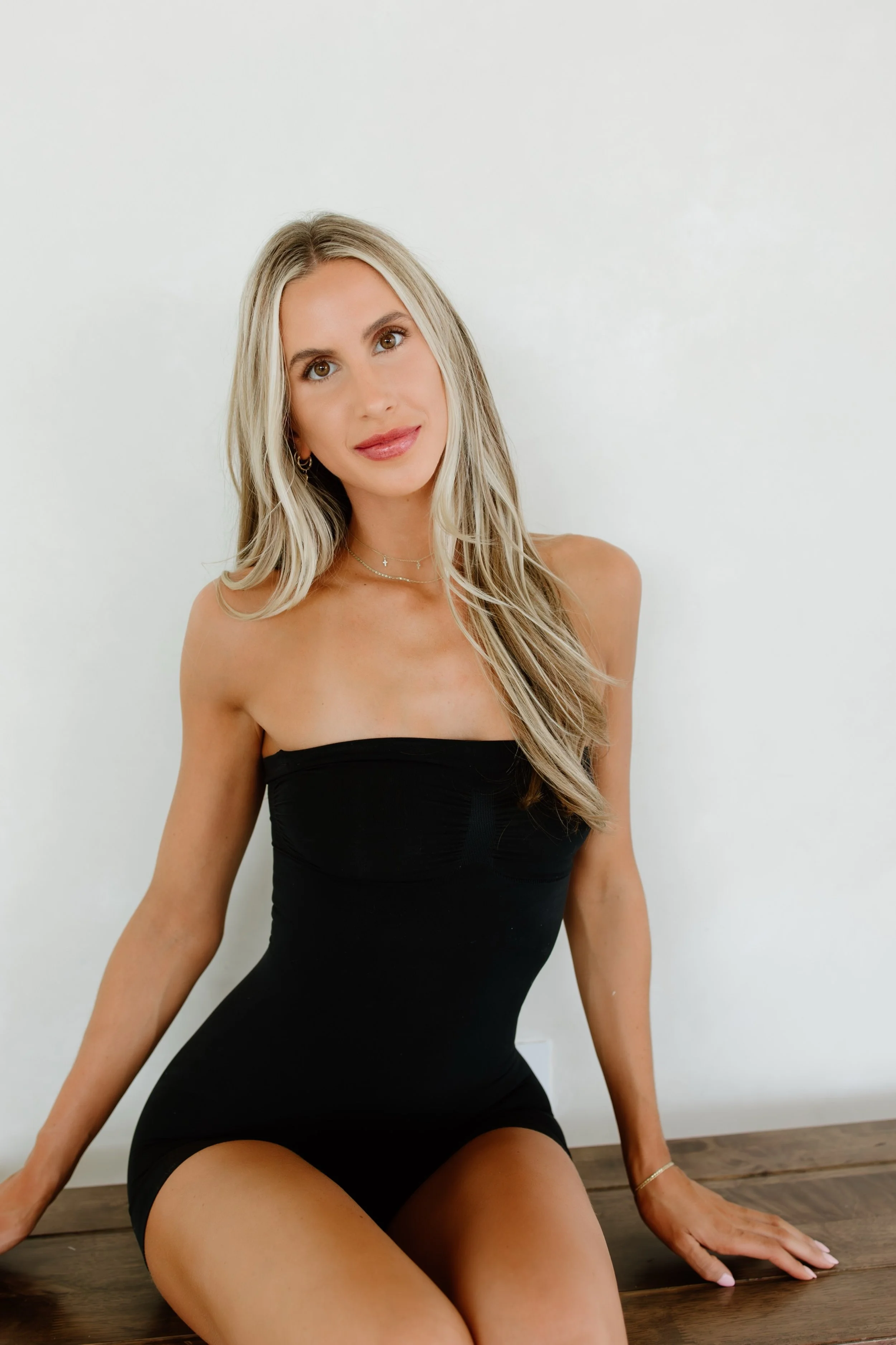 A woman with long blonde hair, wearing a black strapless dress, sitting on a wooden surface against a plain light-colored wall.
