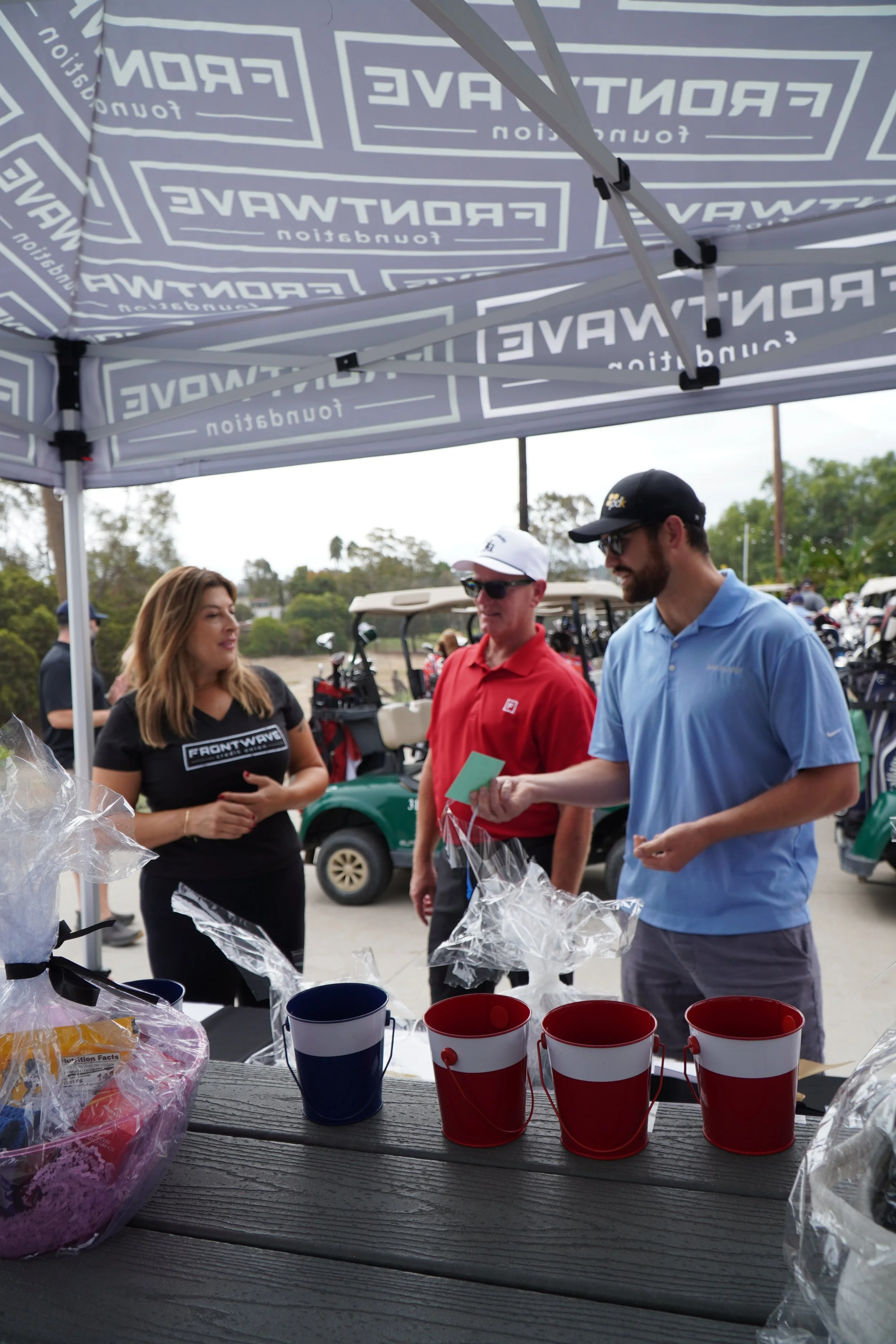 People at a booth under a tent with golf carts and trees in the background, two red cups, a blue cup, and wrapped items on a wooden table in the foreground.