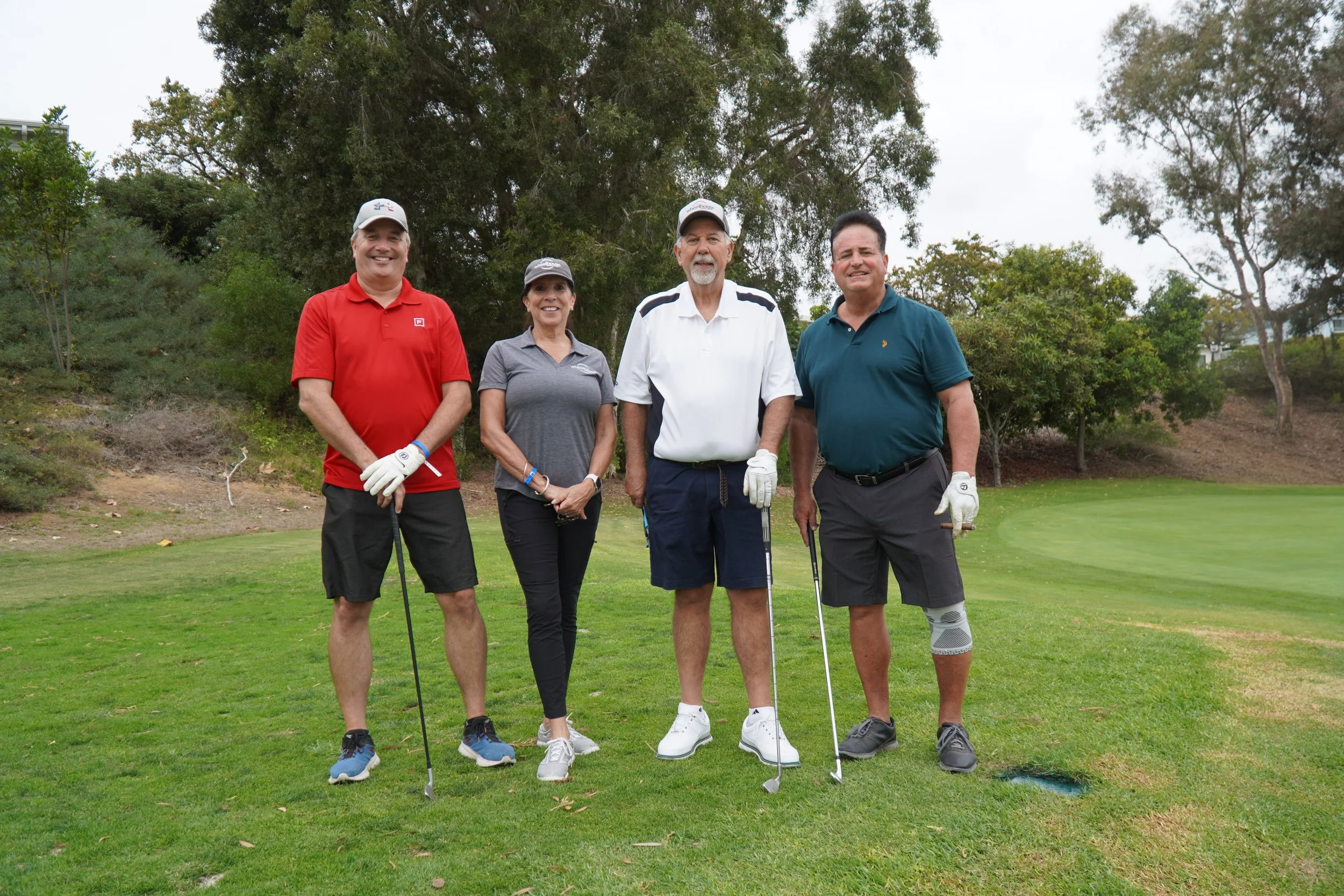 Four people standing on a golf course, smiling at the camera, with trees in the background. They are wearing golf attire and holding golf clubs.