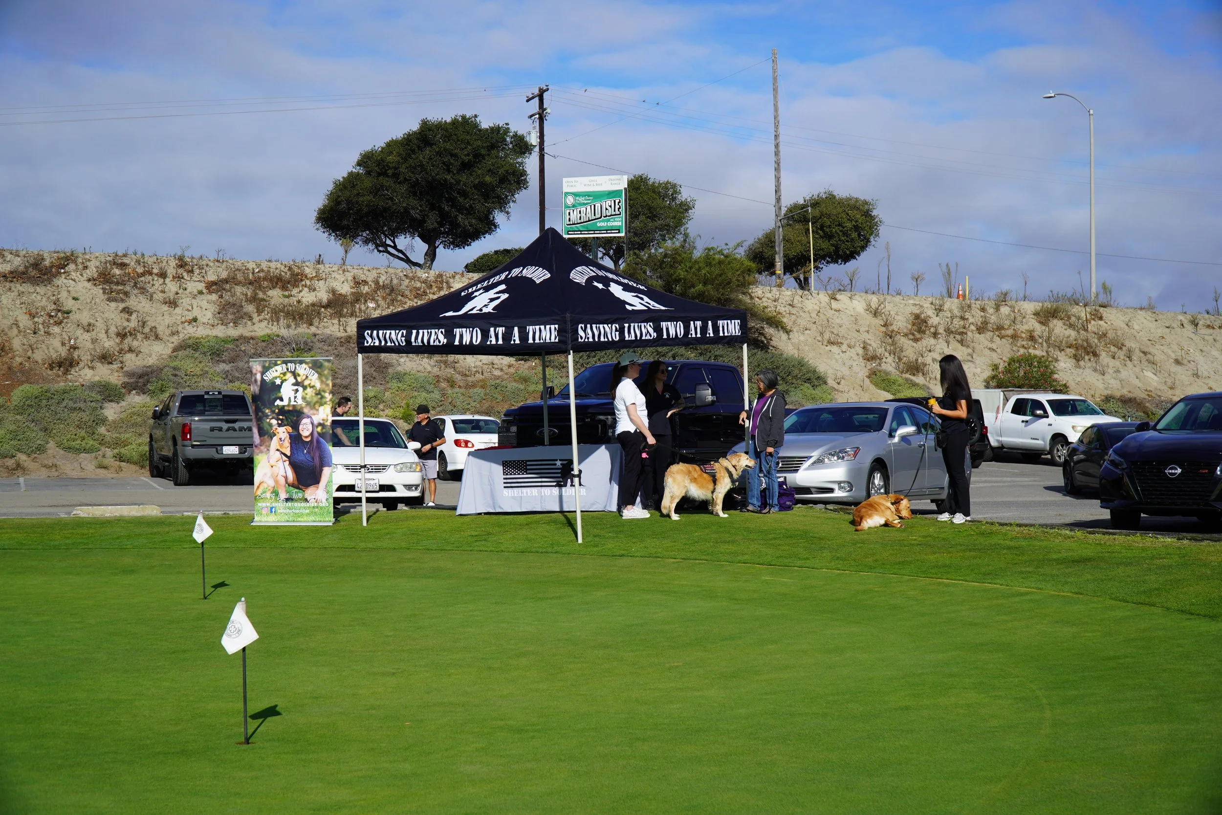 People gathered under a black tent with the slogan 'Saving Lives, Two at a Time,' at a charitable event for animal rescue, with dogs, a table, and cars parked nearby on a sunny day.
