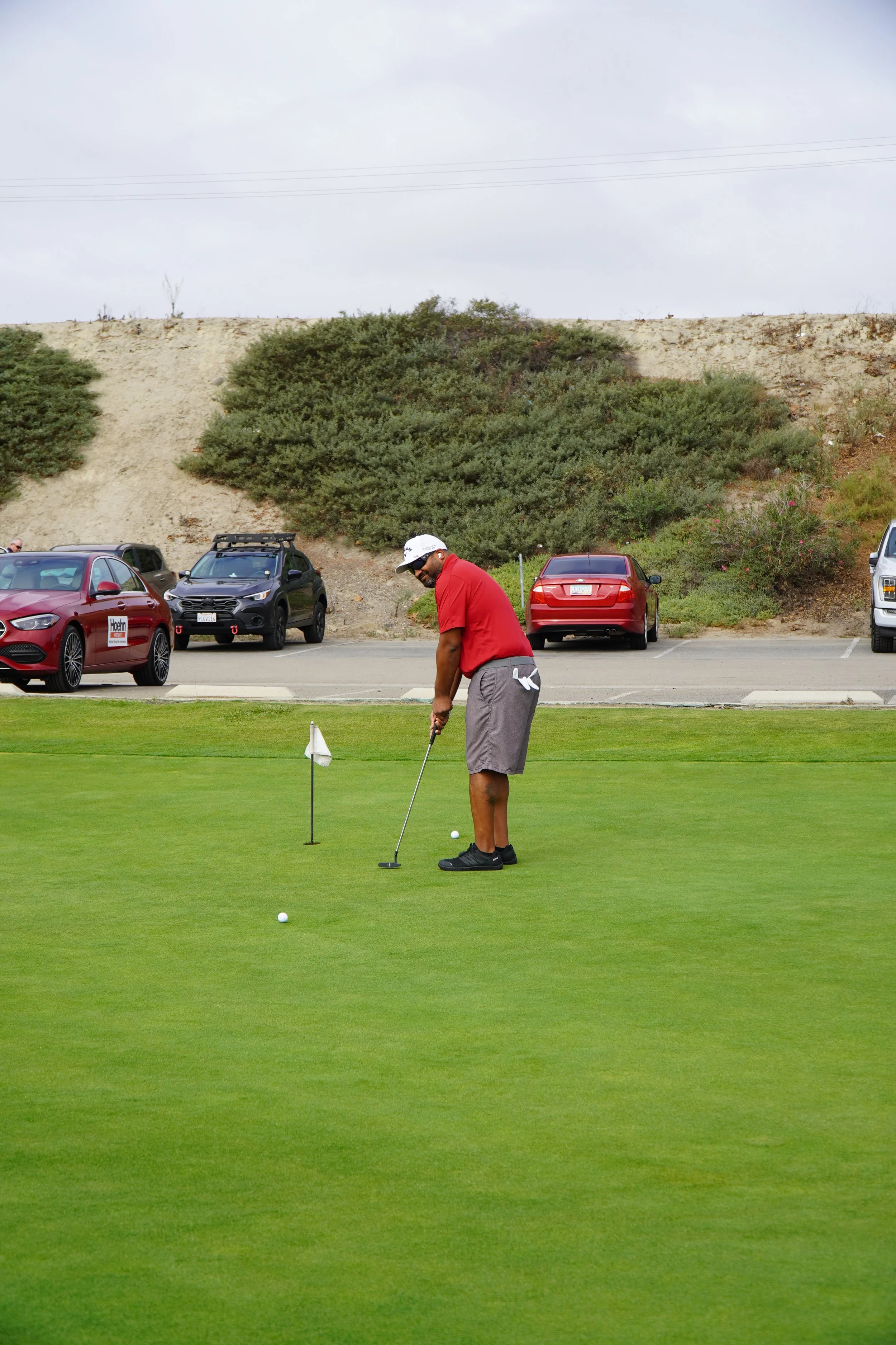 A man in a red shirt, gray shorts, and black shoes preparing to putt a golf ball on the green, with a small flag marking the hole, at a golf course.