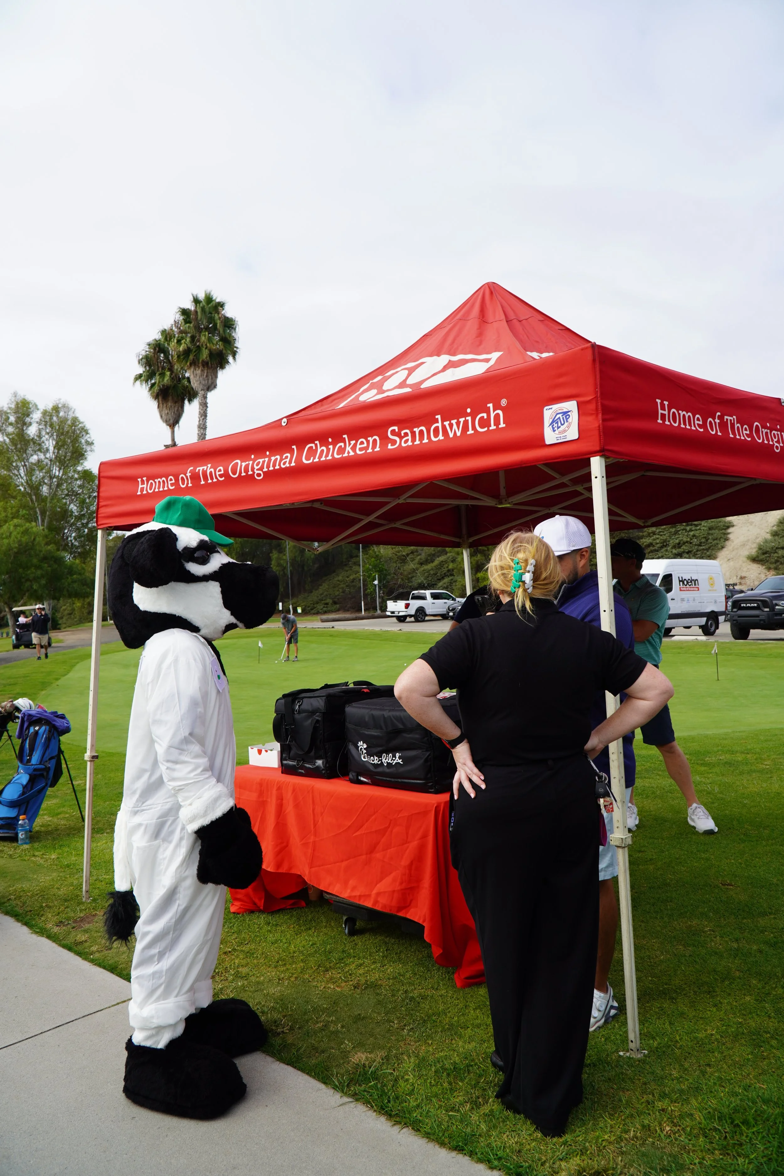A person in a cow mascot costume standing in front of a red tent at a golf course. The tent has the text 'Home of The Original Chicken Sandwich'. Two women and a man are talking under the tent, with golf equipment and vehicles in the background.
