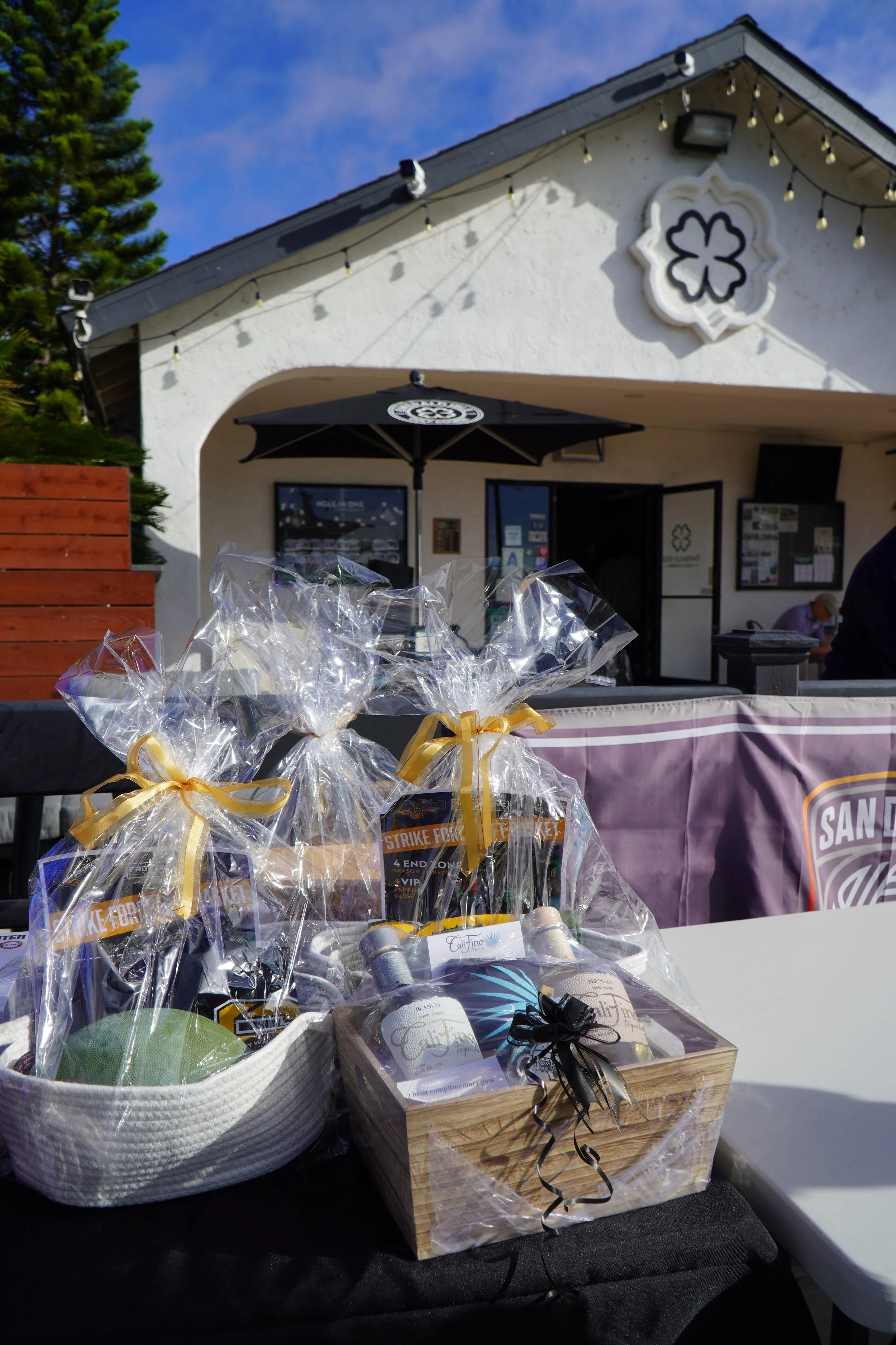 Gift baskets wrapped in clear cellophane with yellow ribbons on a table outside a white building with a black umbrella and string lights.
