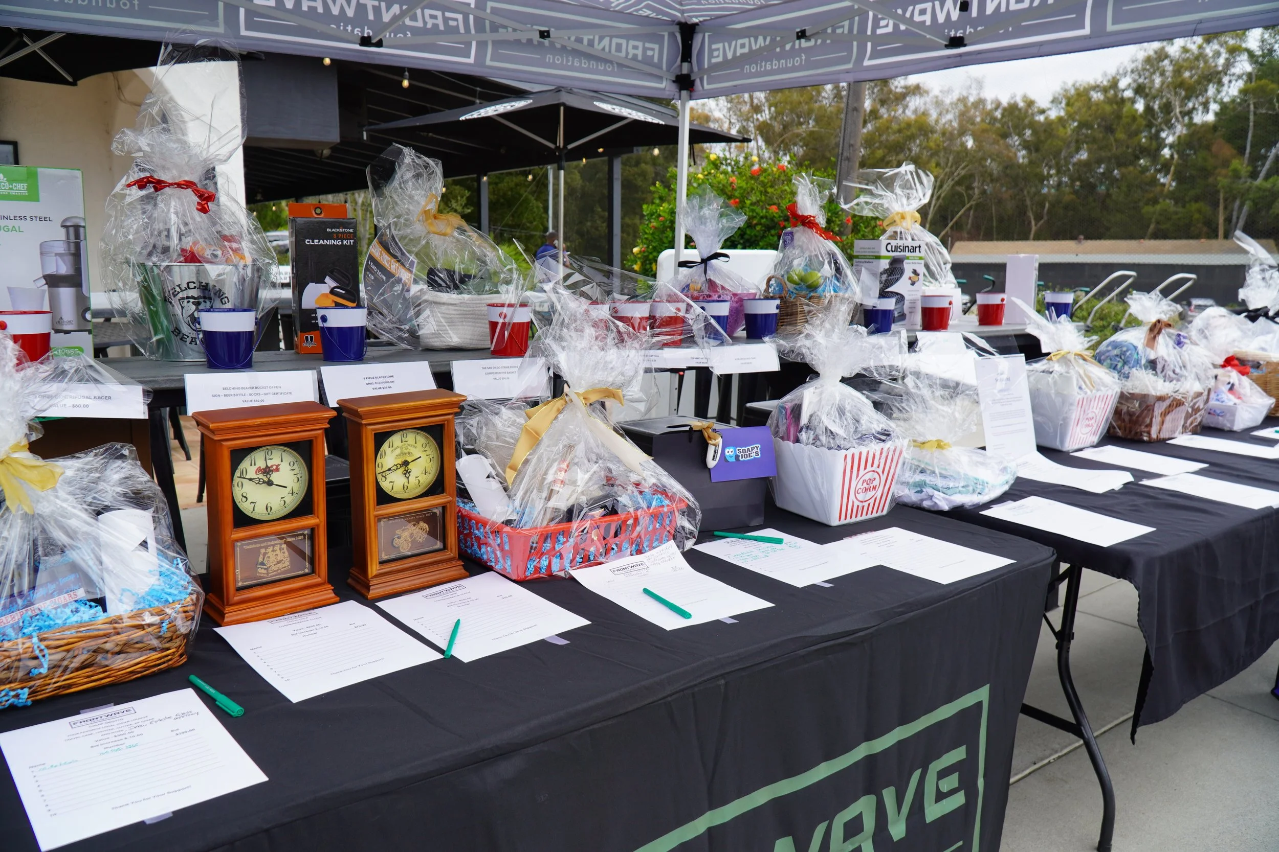 Auction tables set up outdoors with various wrapped gift baskets, clocks, and items for bidding, under tents with people in the background.