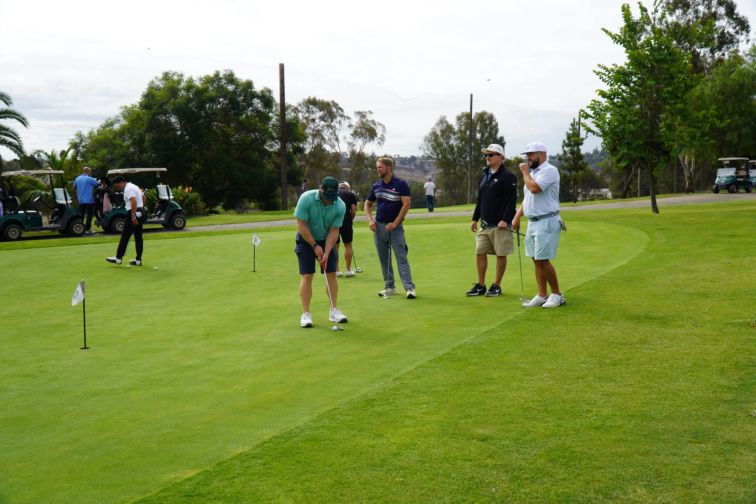 Group of men playing golf on a putting green, with golf carts and trees in the background.