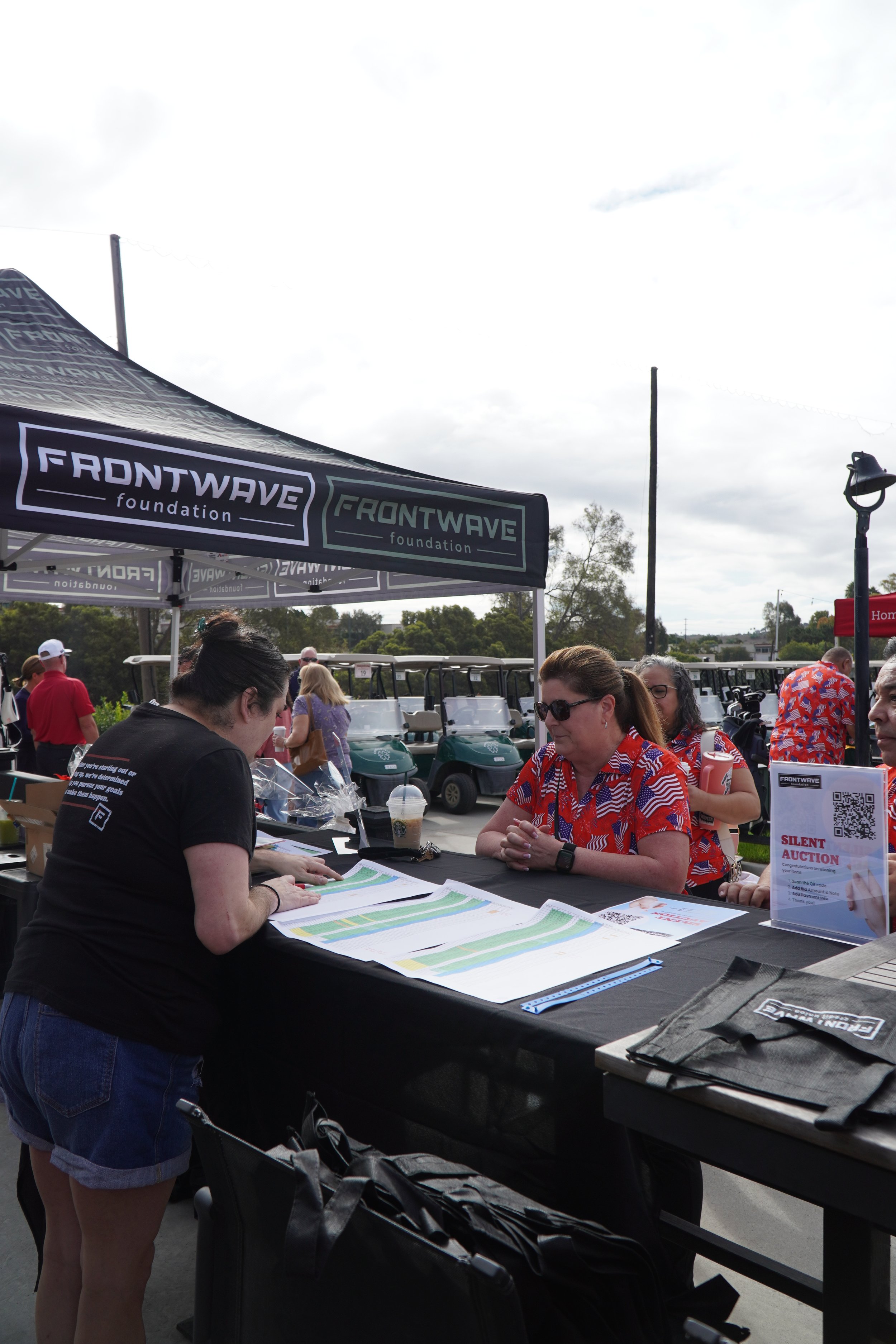 A woman at a registration booth with a woman in sunglasses and a red-patterned shirt, at an outdoor event under a large canopy with "Frontwave Foundation" logo, surrounded by golf carts and other attendees.