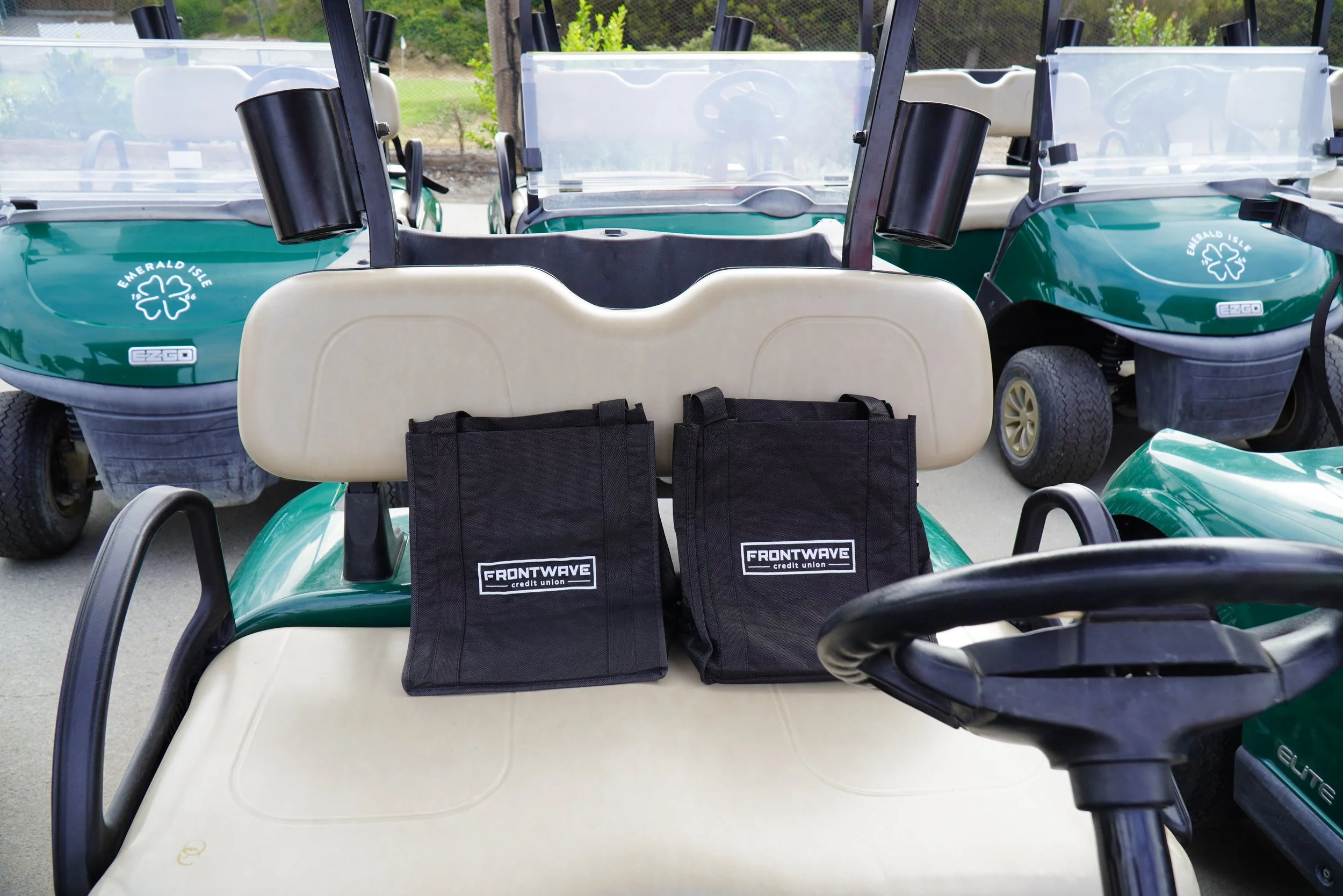 A row of green golf carts parked outdoors, with two black Frontwave credit union bags placed on the beige seat of the central golf cart.