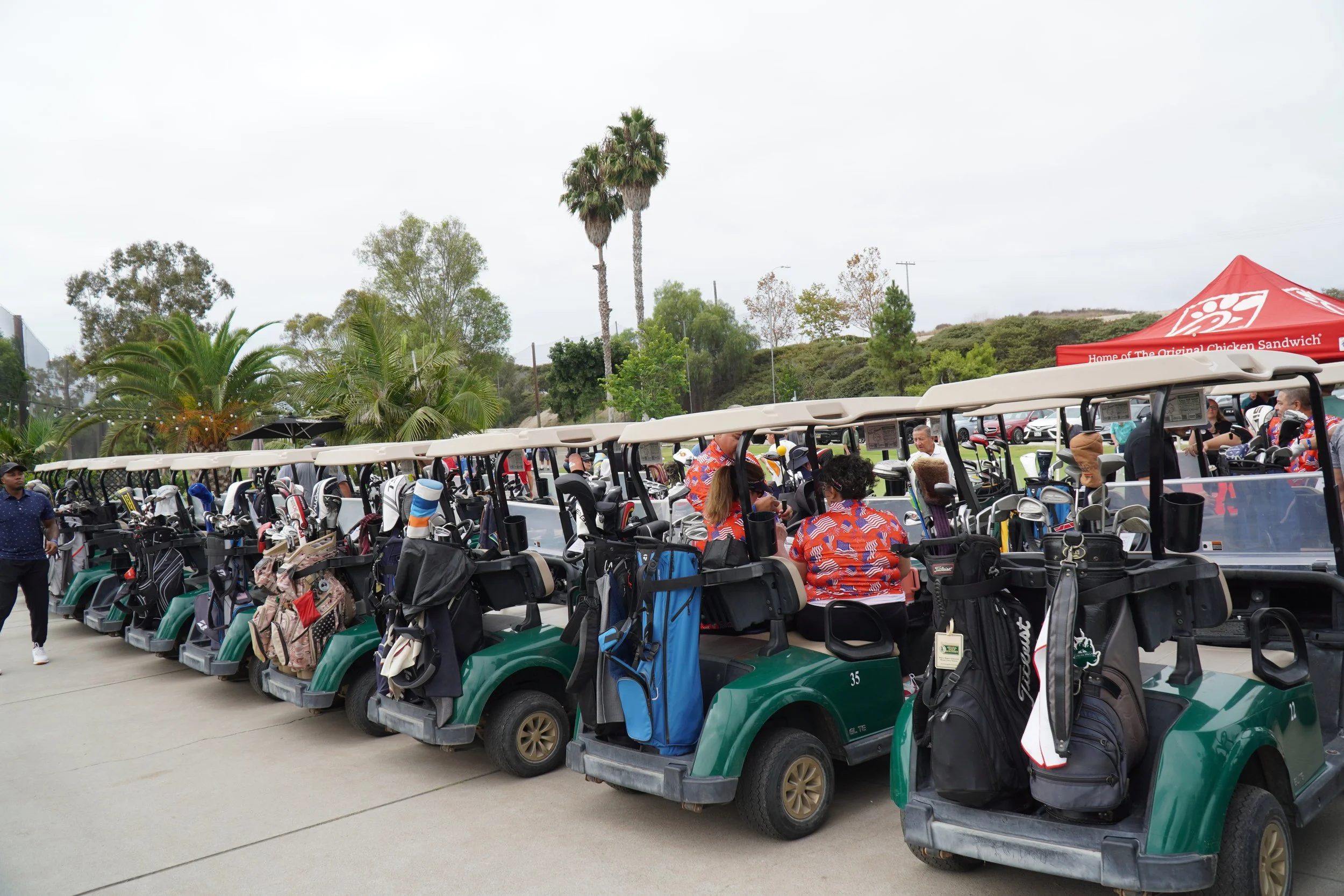Multiple green golf carts with golf bags parked in a row at a golf course, with people sitting inside and standing nearby.
