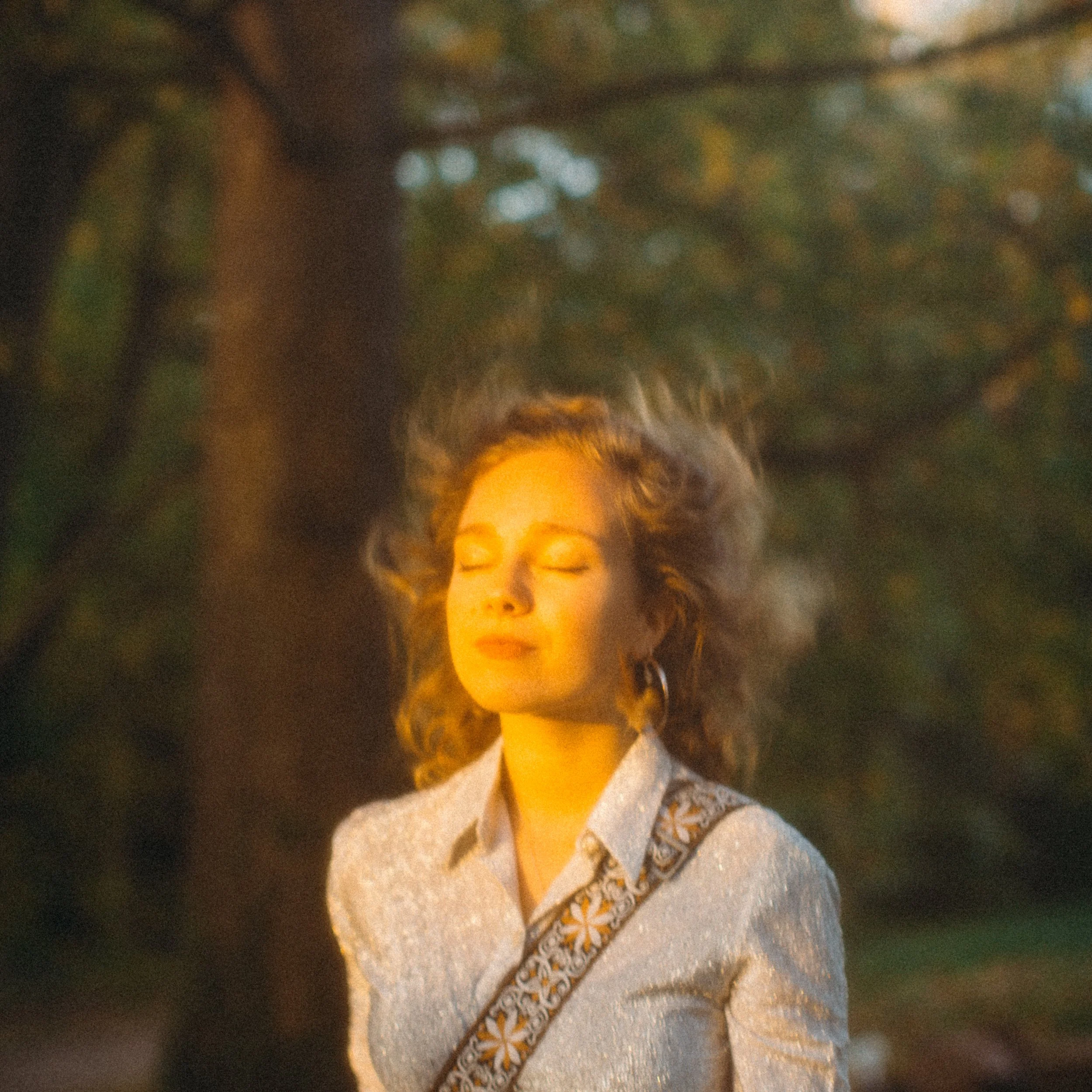 Jeune femme aux cheveux bouclés et aux yeux fermés, portant une chemise claire et un sac à bandoulière avec un motif floral, se tenant devant un arbre avec des feuilles vertes, en plein air, pendant le coucher du soleil.