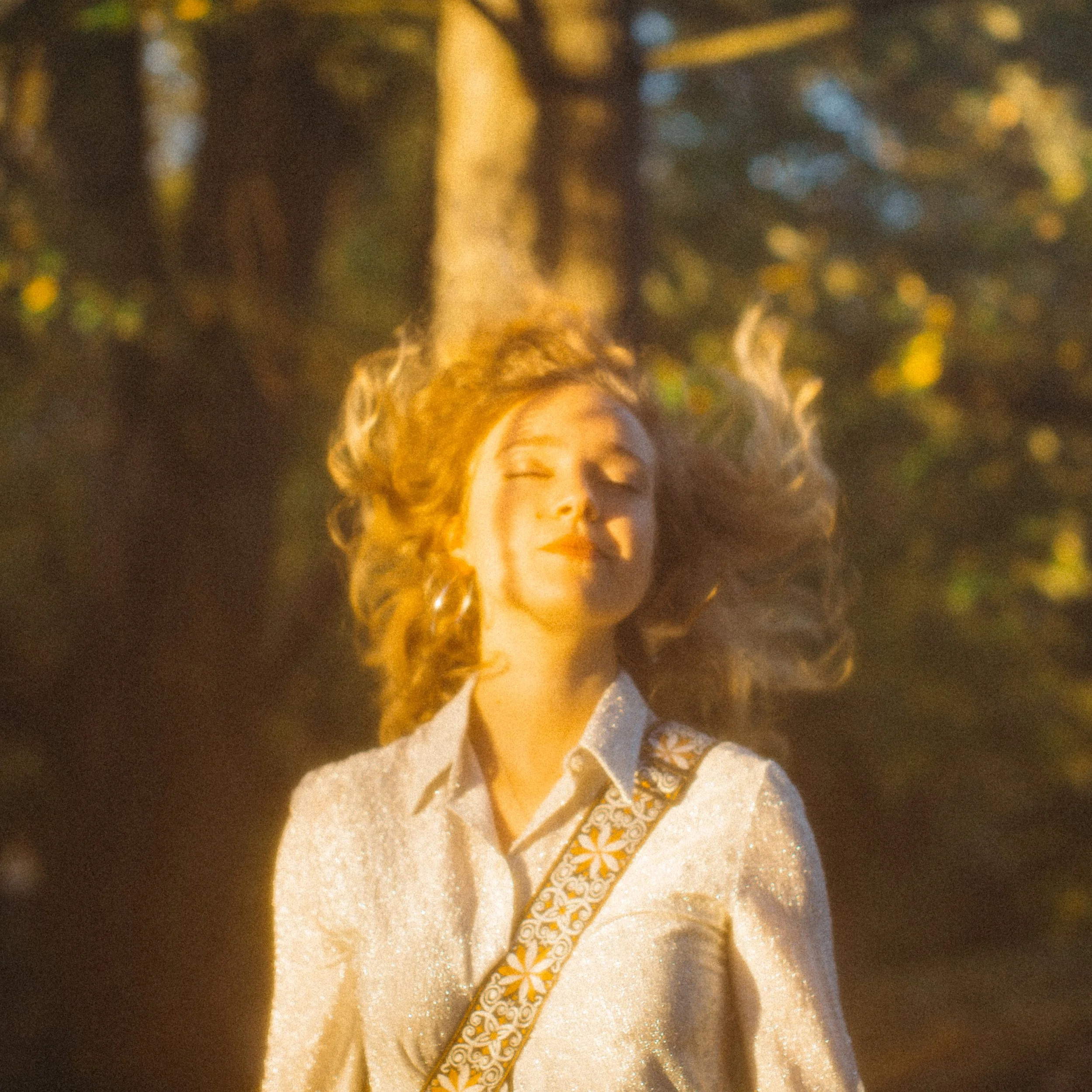 Une femme avec des cheveux bouclés roux, portant une chemise blanche à paillettes et une courroie à motifs floraux, profite du soleil dans un environnement naturel.