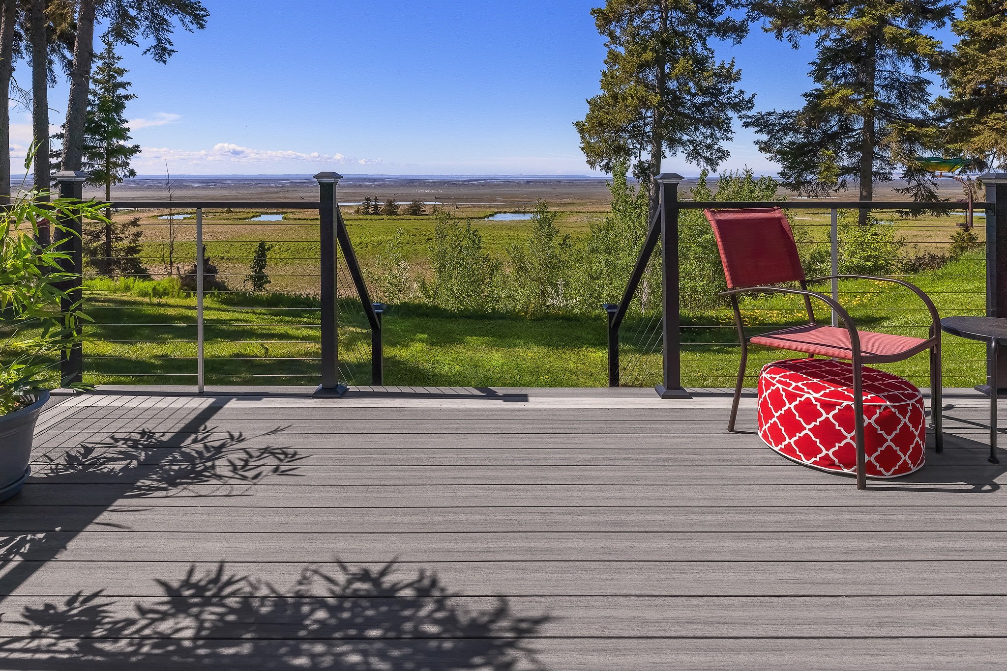 A composite deck with a red chair, a red and white ottoman, and a small black table, overlooking green fields and a distant view of water under a clear blue sky.
