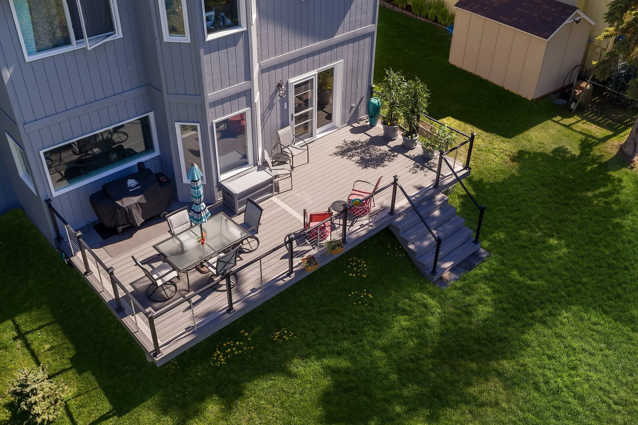 Aerial view of a gray house with a composite deck furnished with outdoor chairs, a dining table with an umbrella, potted plants, and a grill, surrounded by a green lawn with some stepping stones and a small shed in the background.