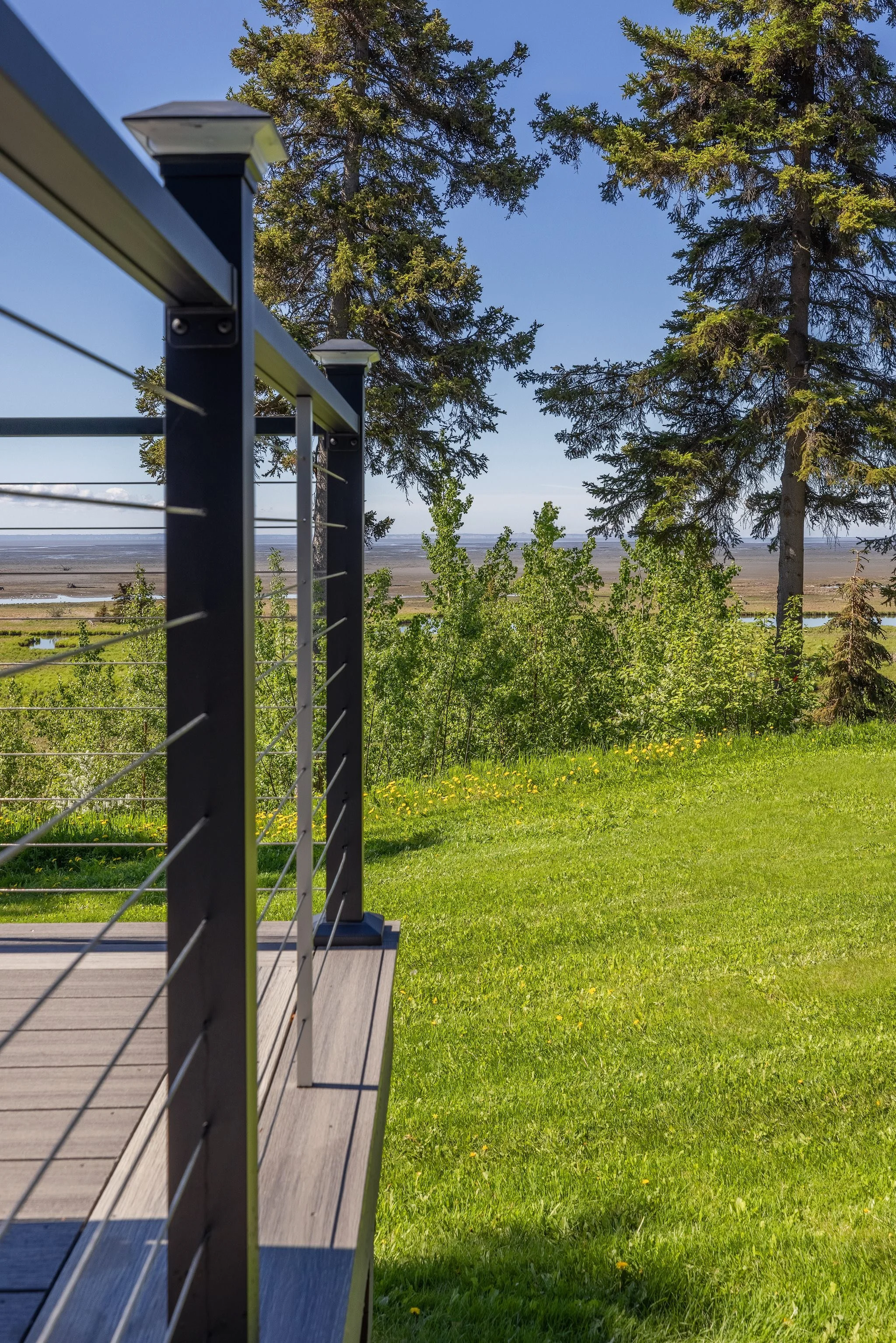 View from a deck with a metal railing overlooking a lush green lawn, trees, and a distant body of water under a clear blue sky.