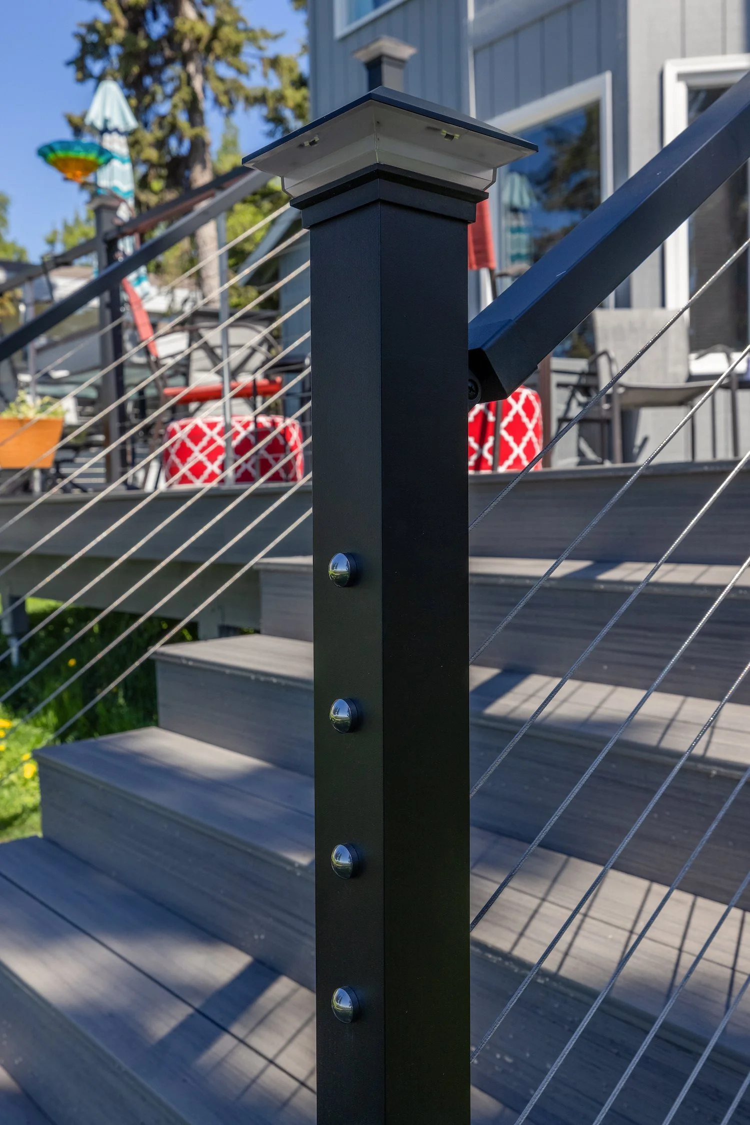 Close-up of black aluminum stair railing post, attached to outdoor stairs with gray wcomposite steps, in front of a house with blue siding and an outdoor patio with red patterned furniture and umbrellas.