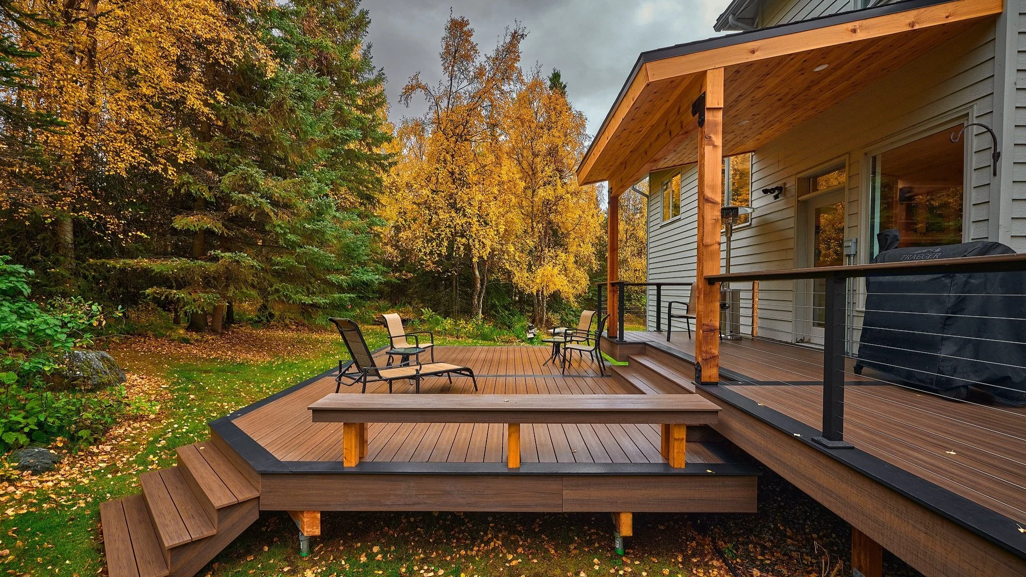 Newly built composite deck outside a house with trees in fall colors, a barbecue grill covered with a black cover, and outdoor chairs, with steps leading down to a grassy yard full of fallen leaves.