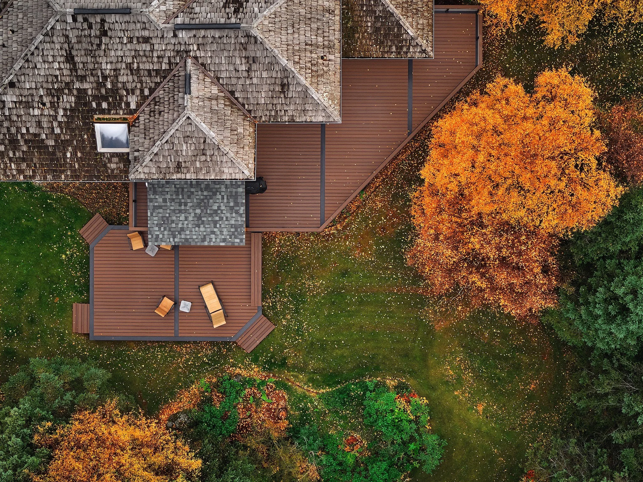 An aerial view of a house with a shingled roof and a composite deck, surrounded by trees with orange and green leaves, indicating autumn.