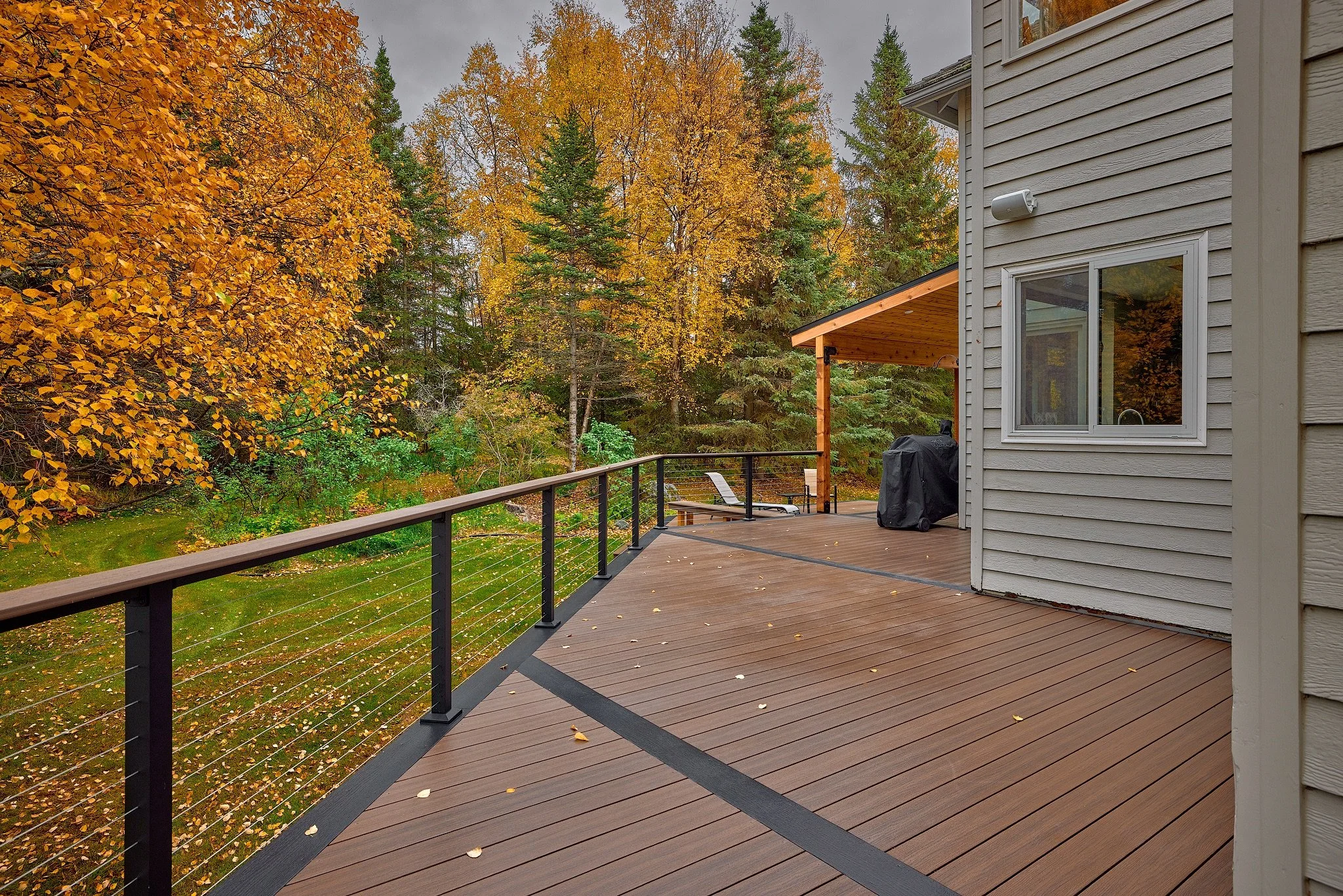 A spacious composite deck attached to a house, overlooking a wooded area with autumn foliage. There is a lounge chair, a barbecue grill, and a small table on the deck.