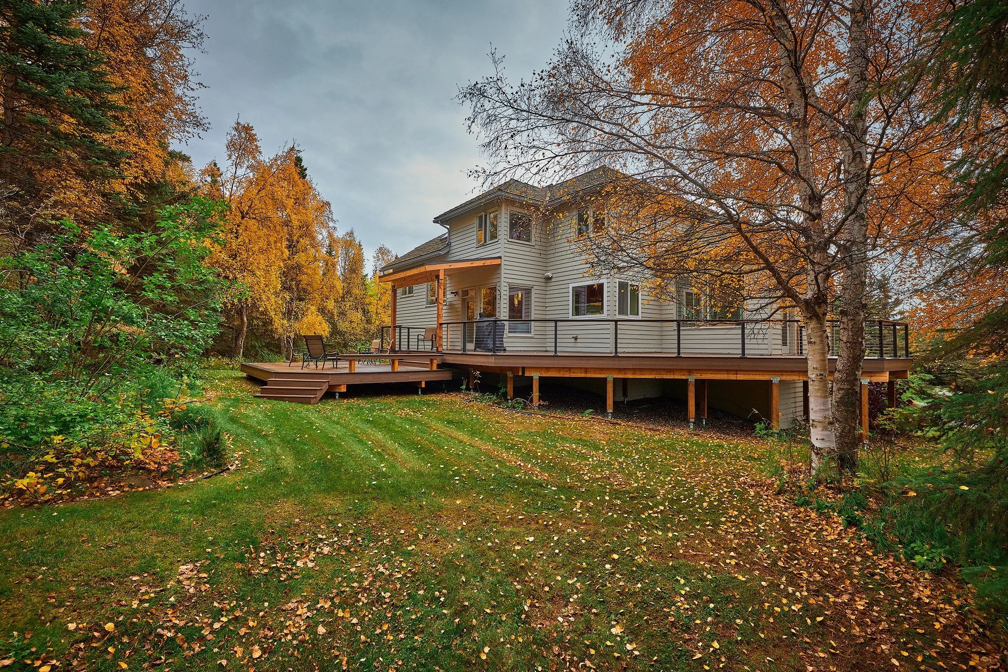 A two-story house with a large composite deck in a forested area during autumn, with fall leaves on the ground and trees with orange and yellow foliage.