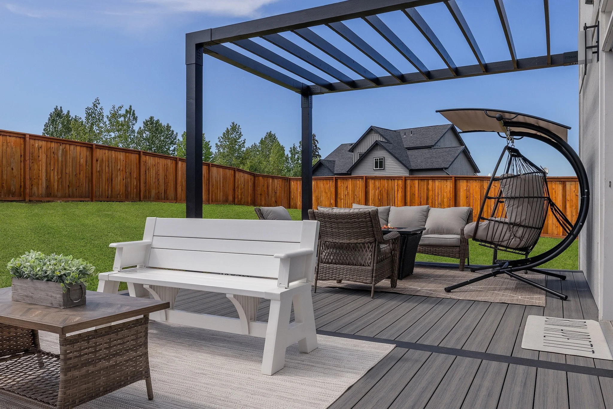 Outdoor patio with white bench, wicker chairs, cushioned sofa, swing chair, and pergola on a composite deck, overlooking a grassy yard with a wooden fence and neighboring house in the background.