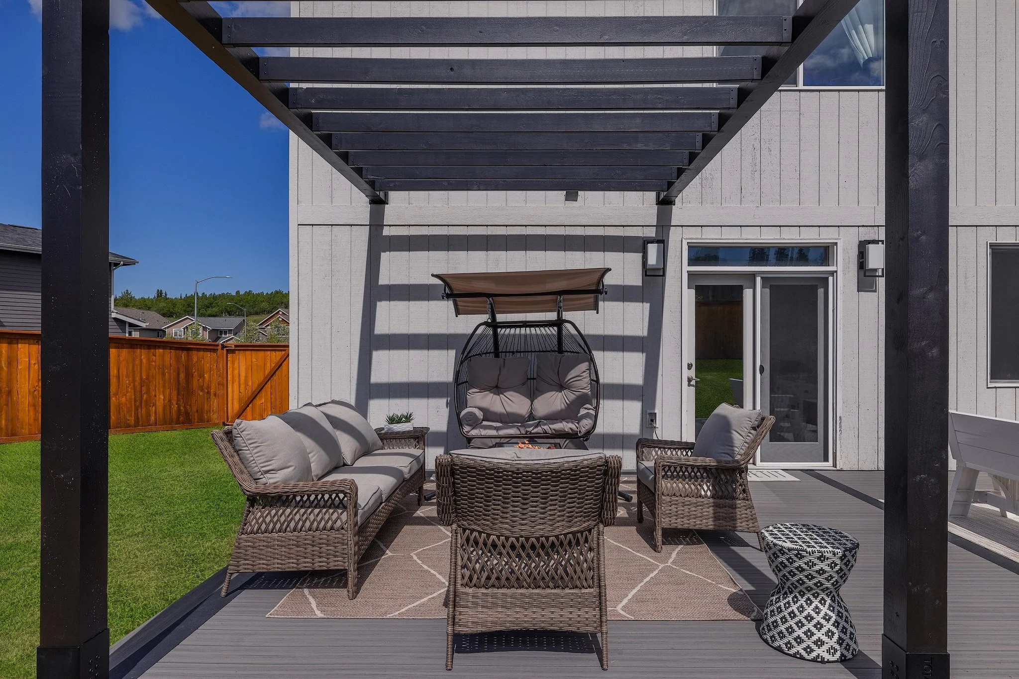 Outdoor patio with wicker furniture, including a sofa, chairs, and a hanging chair with a canopy, and a fire pit on a rug. There is a gray deck, a white house with sliding glass doors, and a wooden fence in the background.